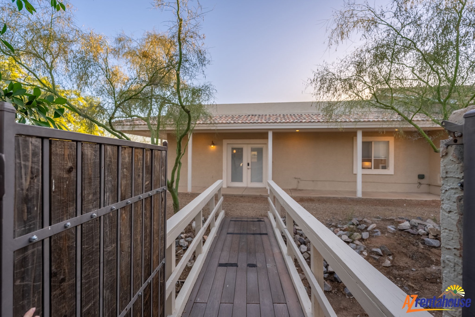 A welcoming single-story home with a charming front walkway, framed by desert landscaping and mature trees. The property features a covered entryway and large windows that invite natural light.