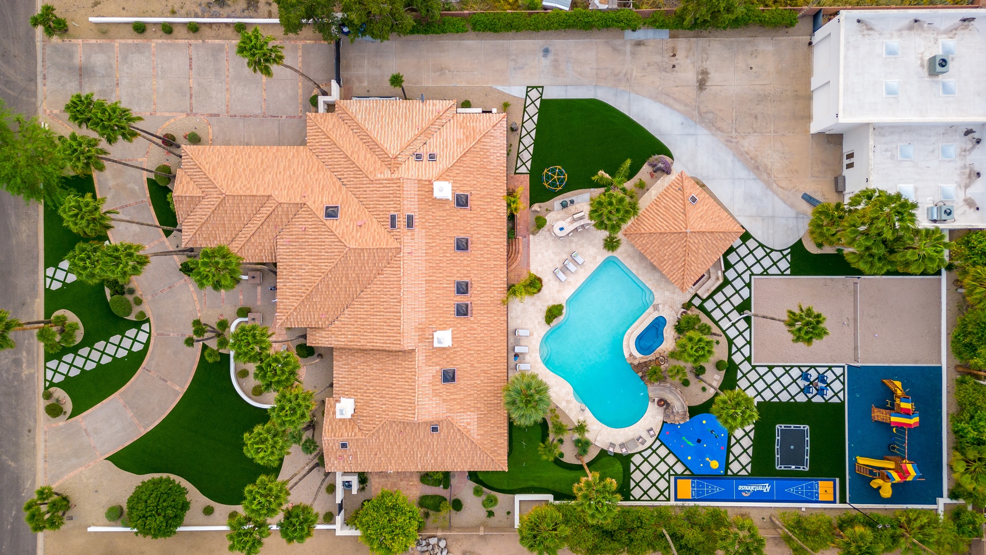 Aerial view of the property featuring a Mediterranean-style building with terra cotta tile roof, private swimming pool, children's play area, and landscaped grounds nestled in a quiet residential neighborhood.