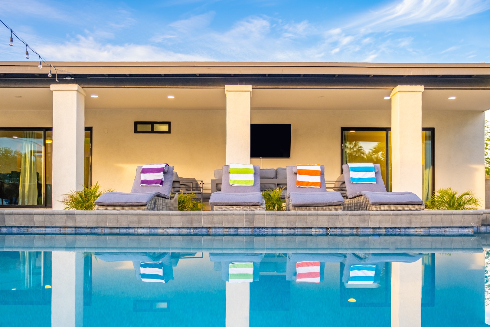Modern pool deck with stylish loungers, an outdoor TV, and pillars framing the sleek design. Perfectly set for sunbathing and poolside relaxation under endless blue skies.