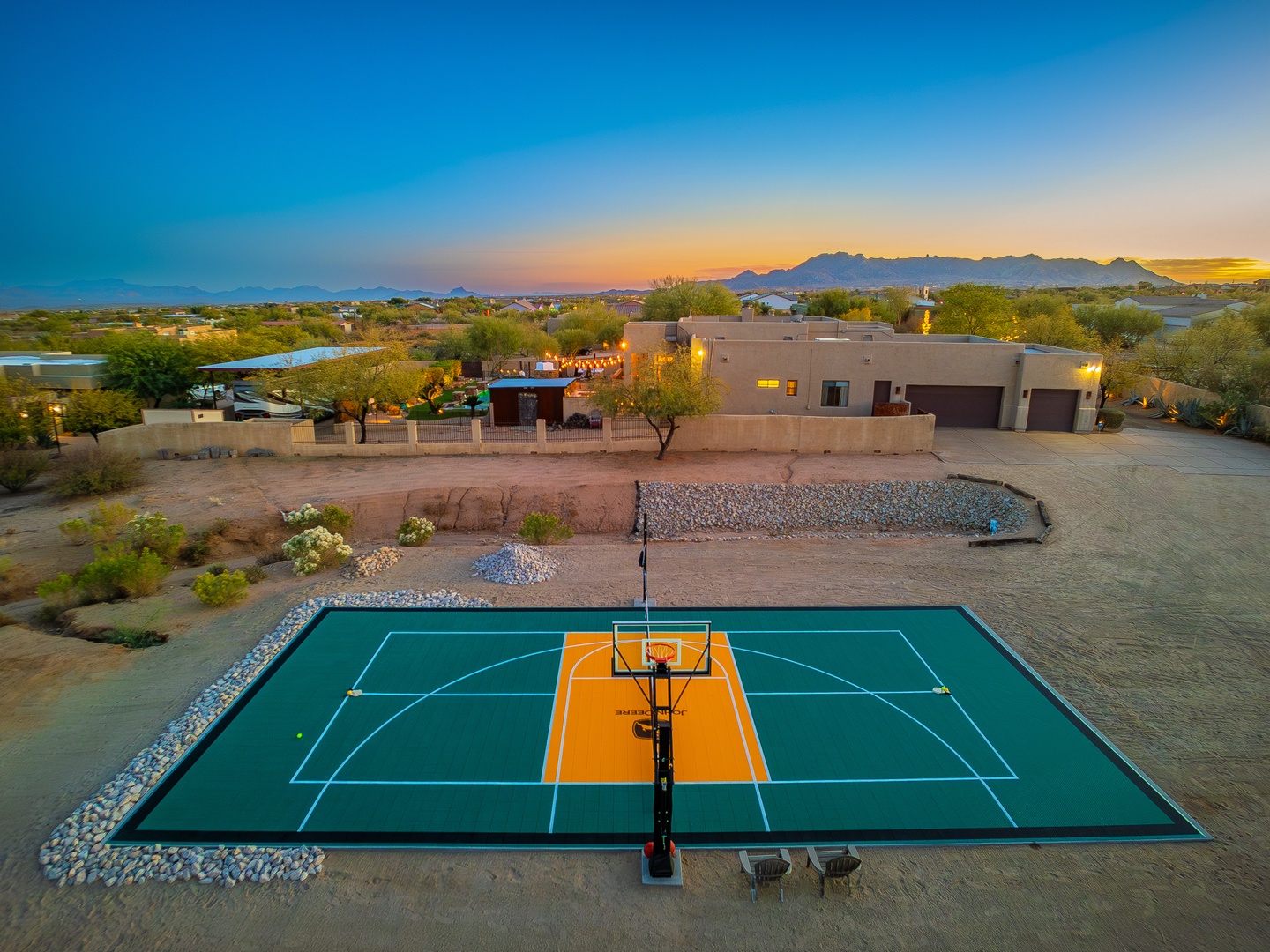 An aerial view captures the property at golden hour, featuring a full-size outdoor basketball court set against desert landscaping and distant mountain ranges, offering guests exceptional recreational amenities.