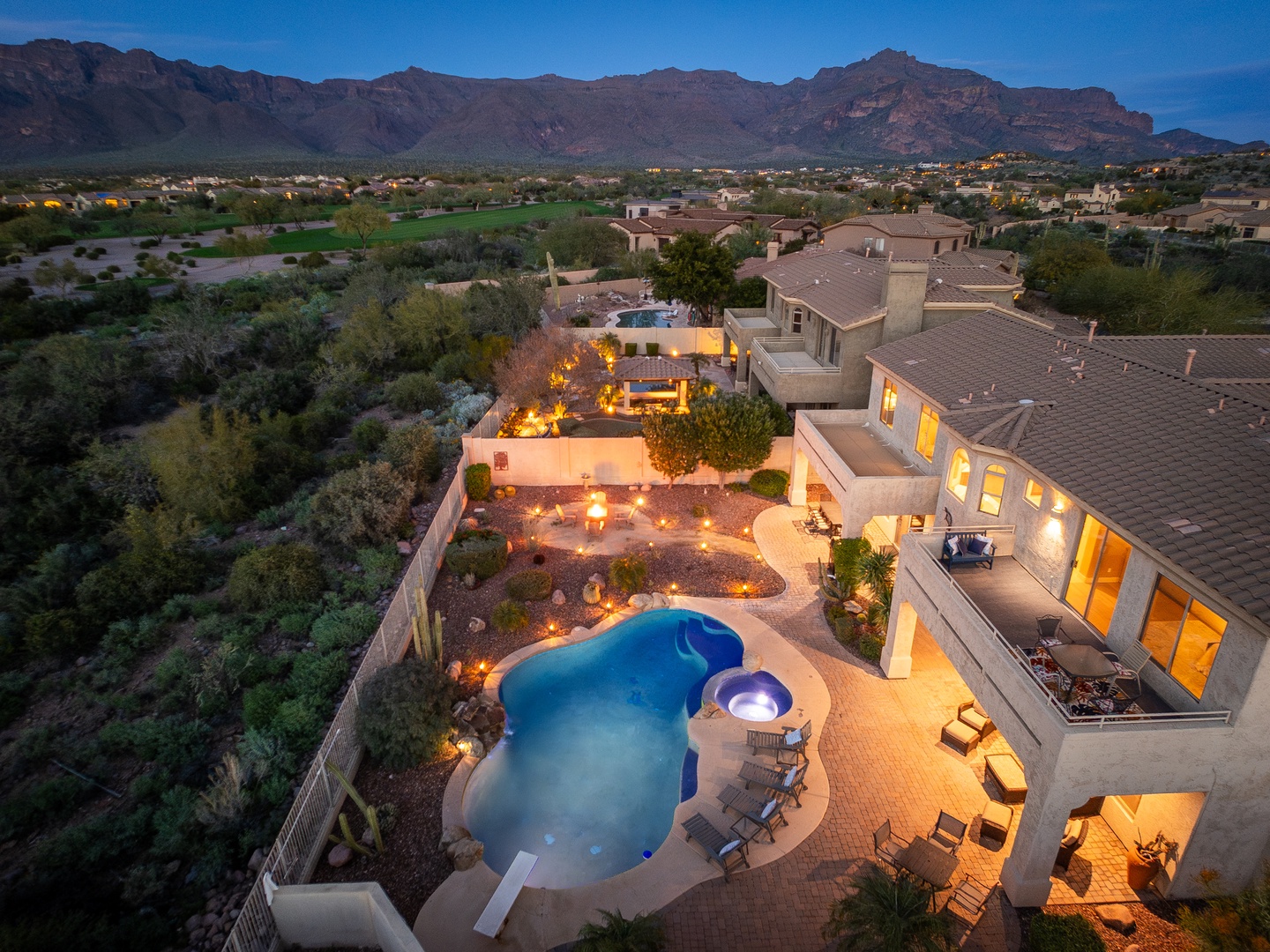 Aerial twilight view showcasing a luxurious desert estate with illuminated pool, spa, and expansive patio, framed by dramatic mountain ranges and lush natural landscape.