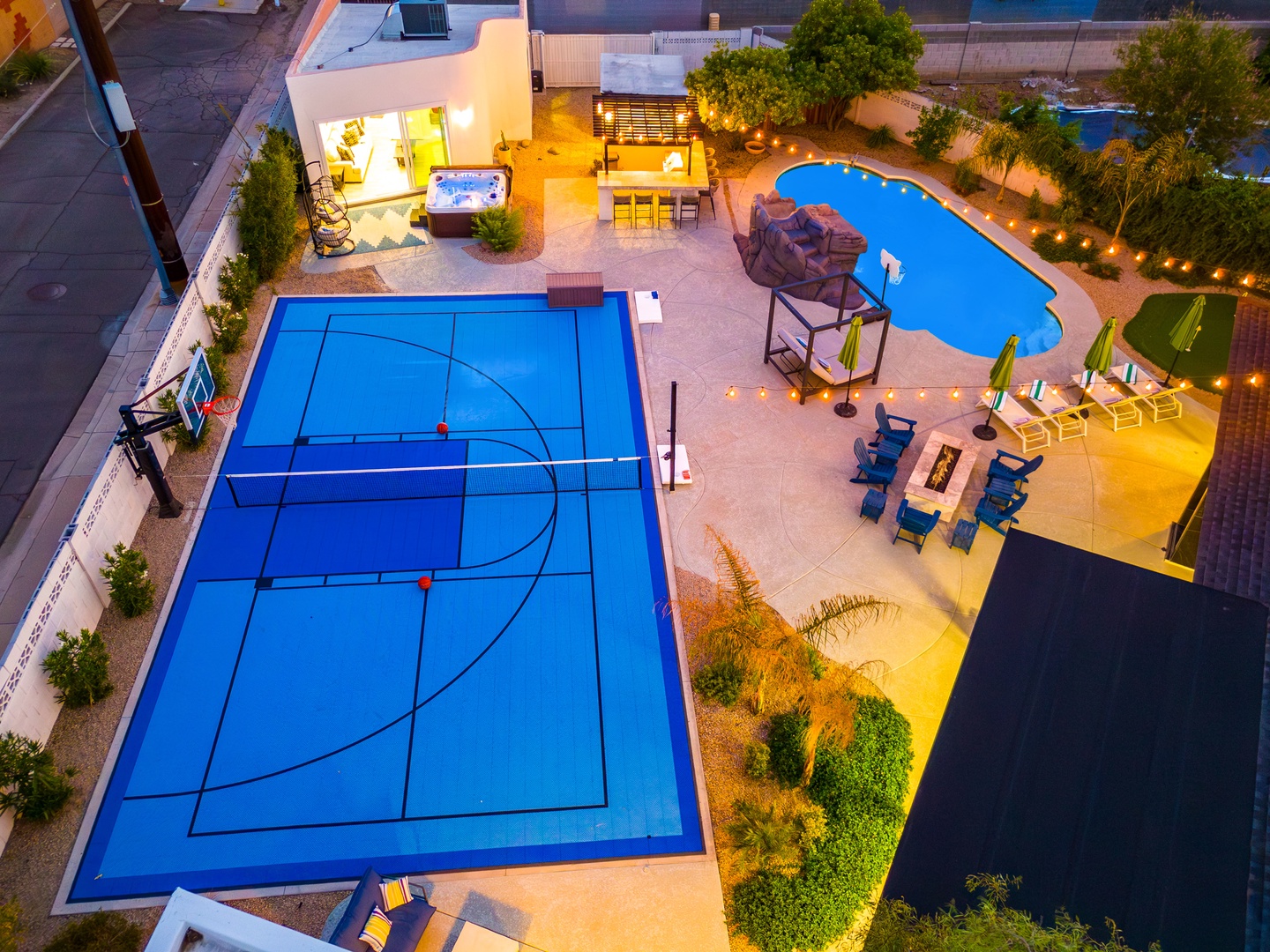 Aerial view capturing an exceptional backyard retreat with a sport court, resort-style pool, hot tub, outdoor dining area, and fire pit—all illuminated under evening lights.