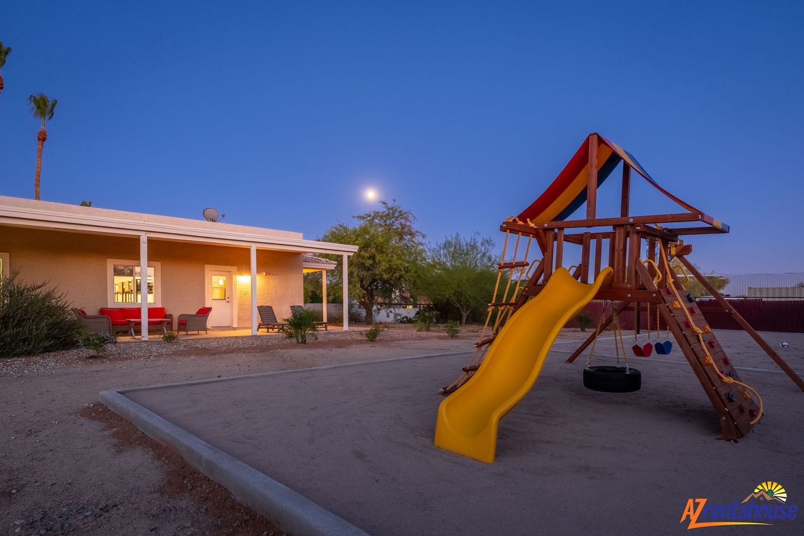 The property features a children's playground with a slide and swings, set against a desert landscape with palm trees and clear evening skies.