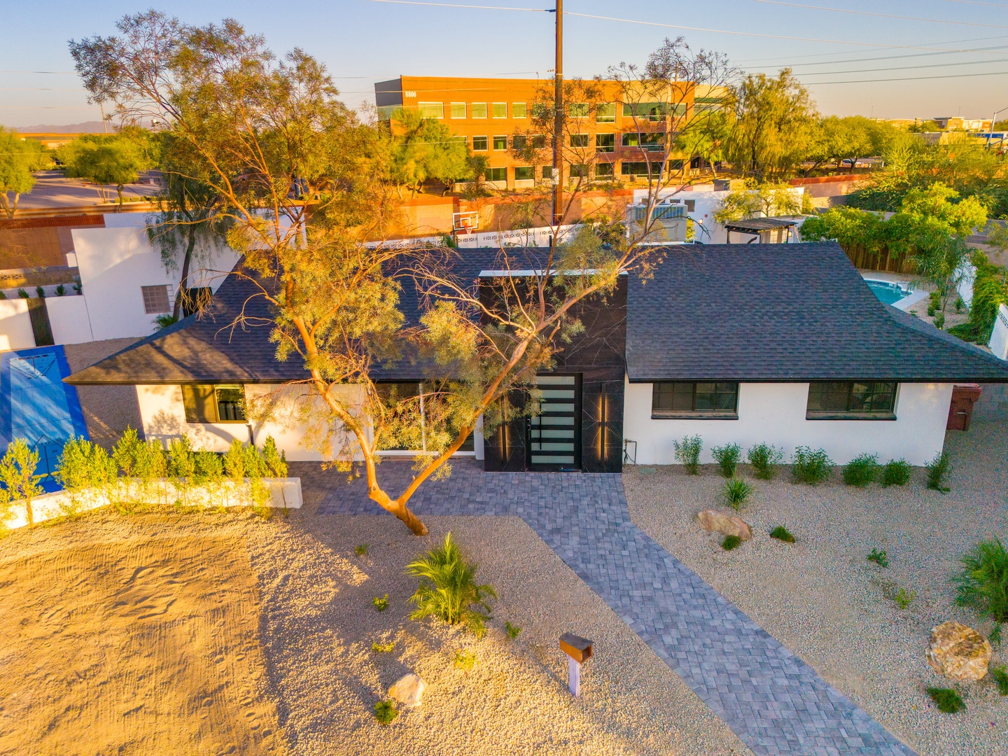 Aerial view capturing the property's modern architecture nestled in a desert landscape, with mature trees, desert landscaping, and surrounding neighborhood structures bathed in warm golden light.