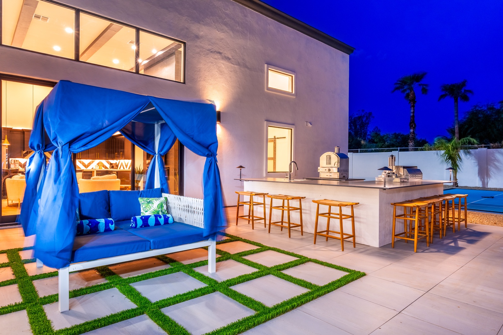 Modern two-story property with illuminated outdoor bar, cabana seating, and palm trees framing the twilight sky at arrival.