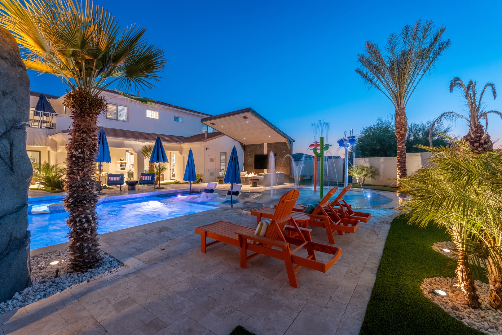 A resort-style backyard oasis at twilight features a sparkling pool, hot tub, colorful splash pad, and elegant covered patio—perfect for creating unforgettable vacation memories under palm trees and blue desert skies.