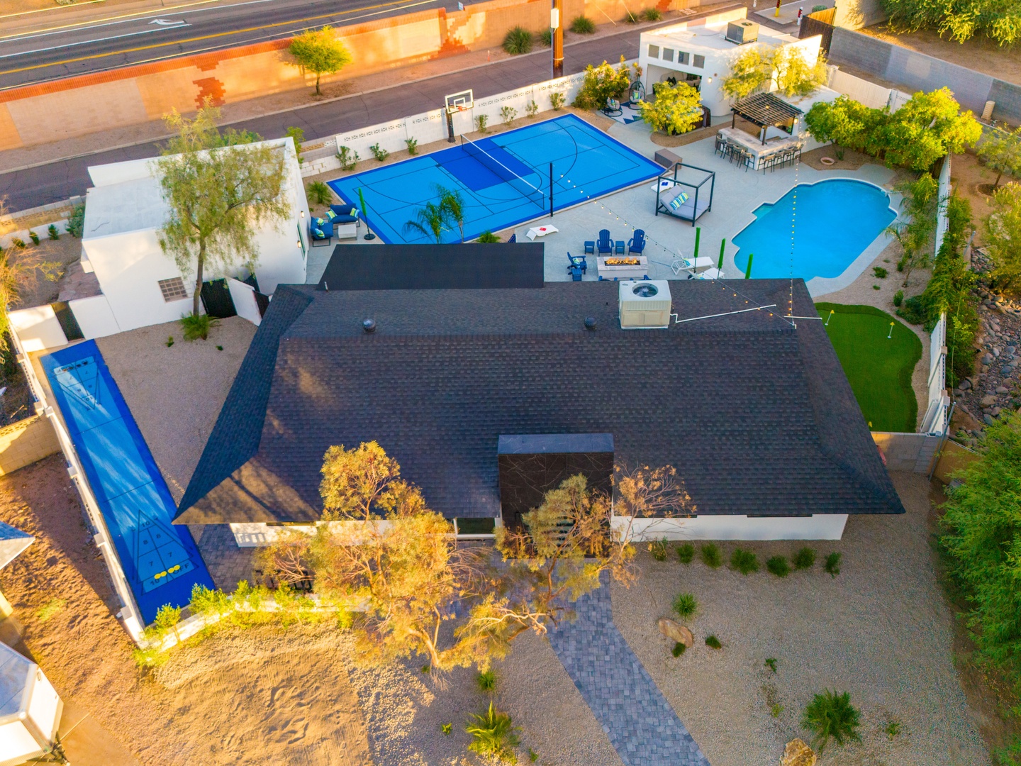 Aerial view of the property showcasing an expansive backyard with a sparkling pool, sport court, outdoor dining area, and desert landscaping with mature trees.