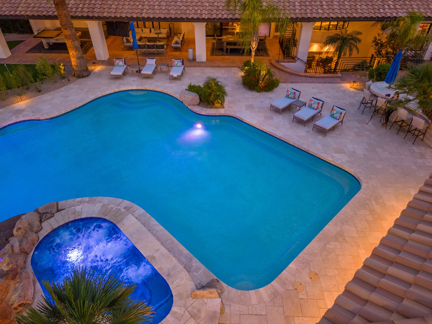 Aerial view of the illuminated pool and hot tub with patio loungers, captured during the warm evening glow as lights create a welcoming ambiance around the courtyard.