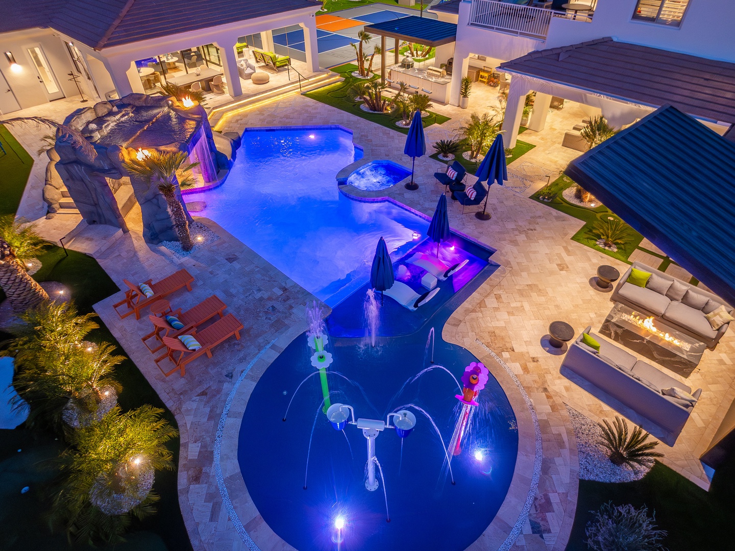 An aerial view showcasing the property's illuminated evening ambiance, featuring a resort-style pool complex with waterslides, hot tub, sunbathing areas, tropical landscaping, and multiple outdoor lounging spaces.