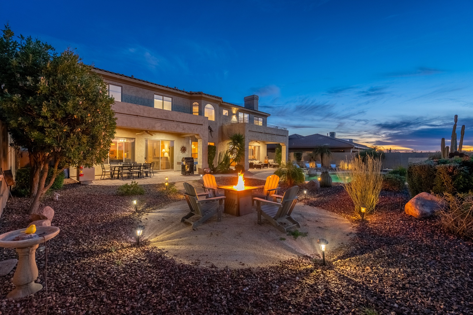 A stunning desert estate at twilight, featuring a spacious backyard with fire pit, multiple seating areas, and striking mountain views framed by native cacti and landscaping.