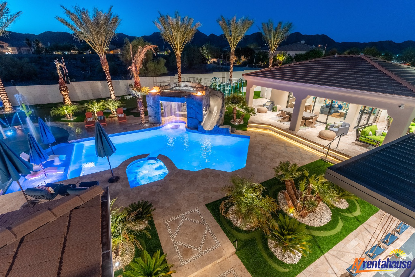 Stunning desert oasis property featuring illuminated palm trees, custom pool with slide, and outdoor entertaining spaces at twilight.