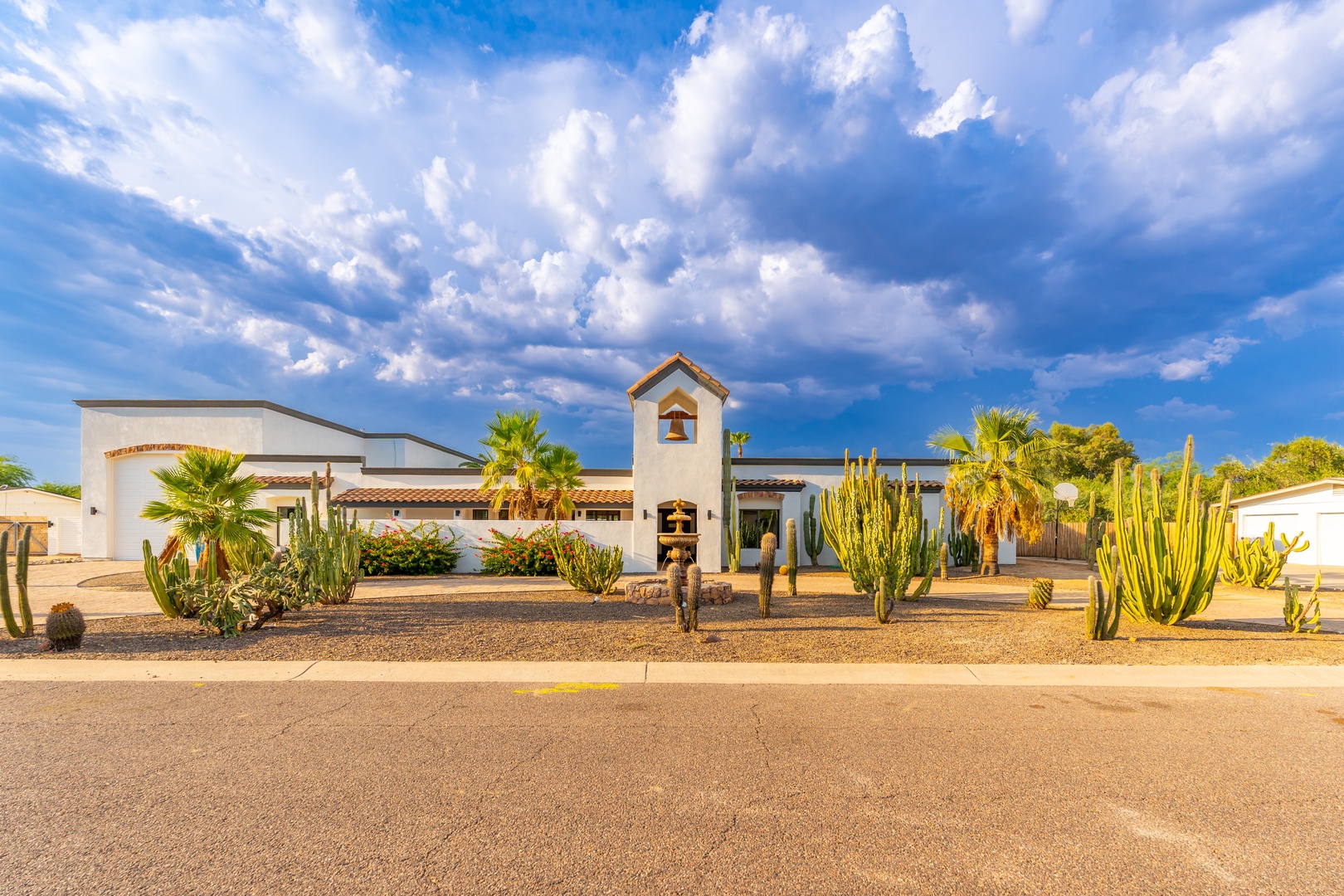 A striking Spanish-style property framed by majestic cacti and palm trees, with a distinctive tower entrance and desert landscaping set beneath dramatic southwestern skies.