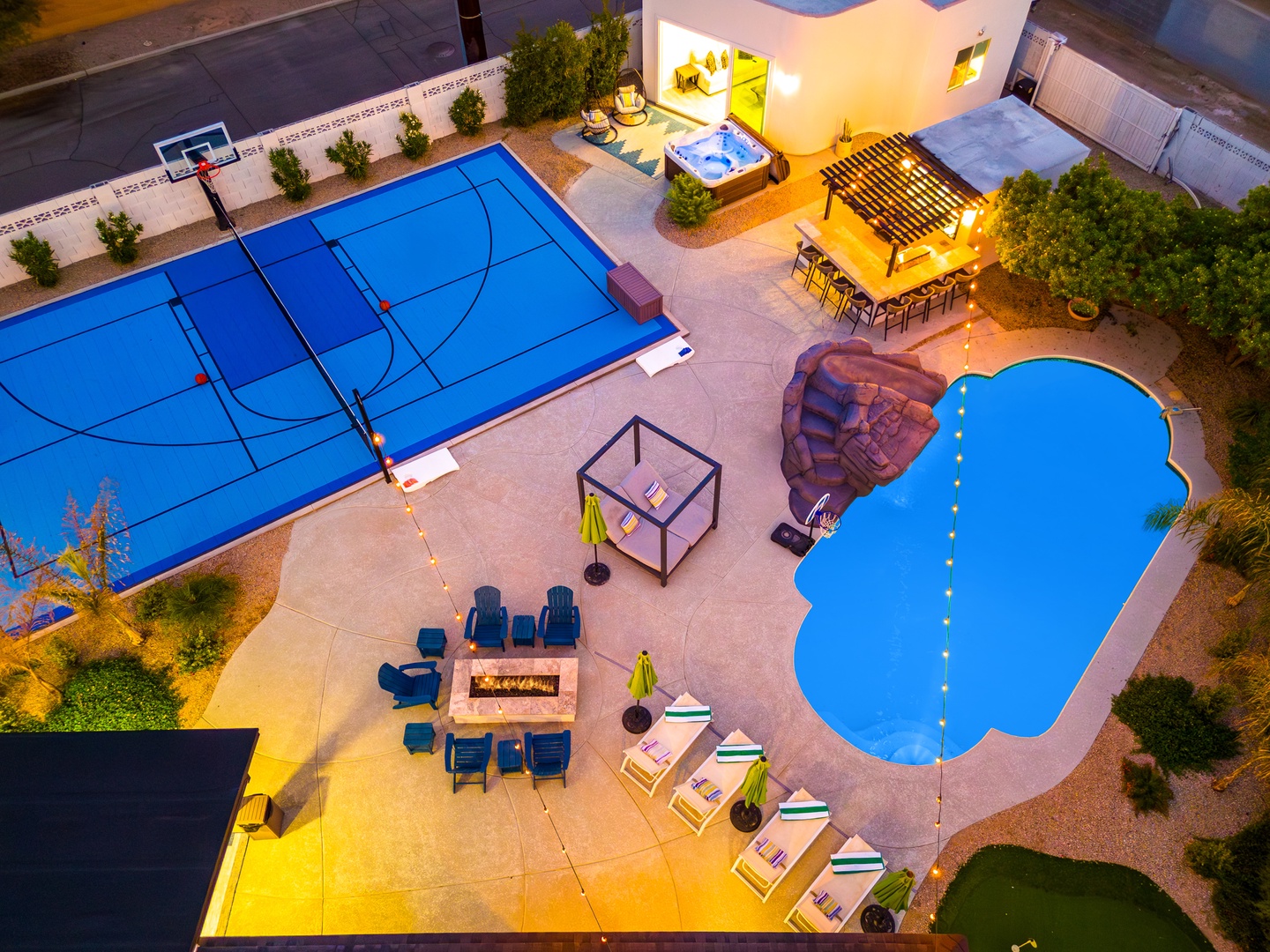 Aerial view of a luxury vacation home featuring full-sized sports court, resort-style pool with waterslide, hot tub, outdoor dining, fire pit seating, and professionally landscaped grounds.