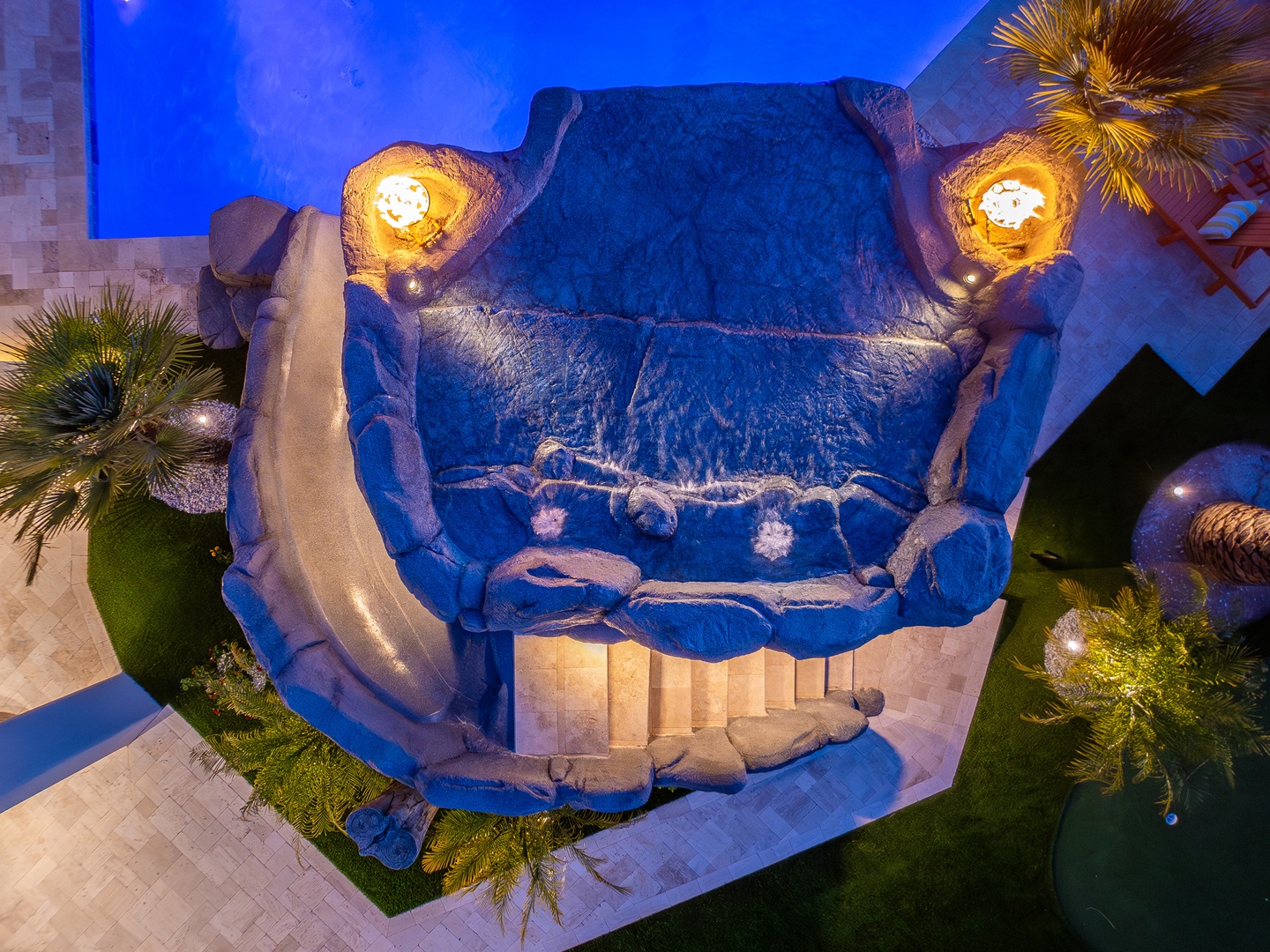 An aerial view showcases the property's distinctive free-form pool surrounded by lush palms, illuminated during twilight hours.