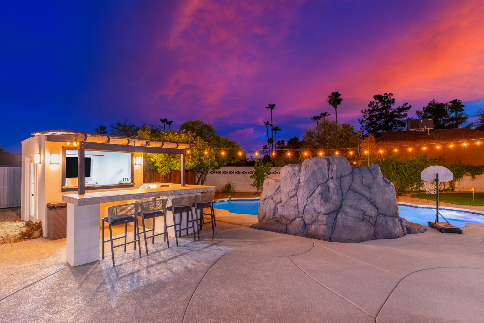 This desert oasis showcases dramatic twilight colors above a modern outdoor entertainment space with an illuminated pool, boulder feature, and covered bar area—ideal for memorable evenings in a uniquely designed backyard retreat.