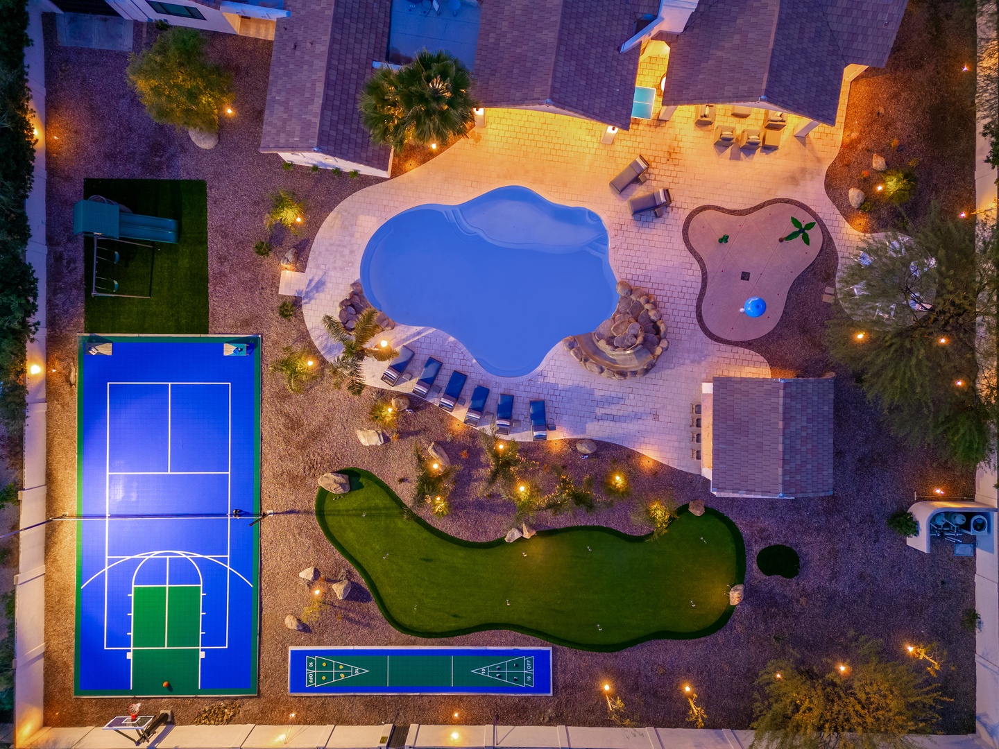 Aerial view of the property showcasing a vibrant outdoor recreation area with a sparkling pool, putting green, tennis court, and children's splash pad, all beautifully lit for evening enjoyment.