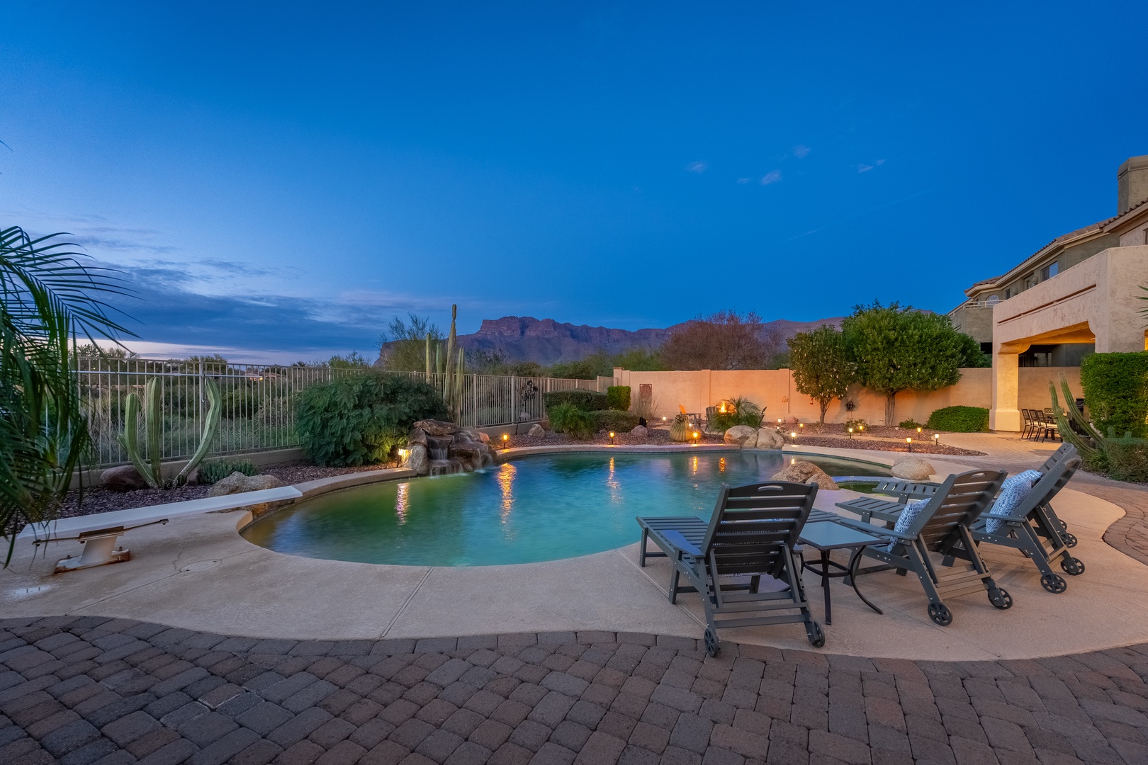 This pool area features dramatic mountain views as a backdrop, surrounded by desert landscaping and lounge seating. The property's exterior showcases southwestern architecture with ambient lighting.