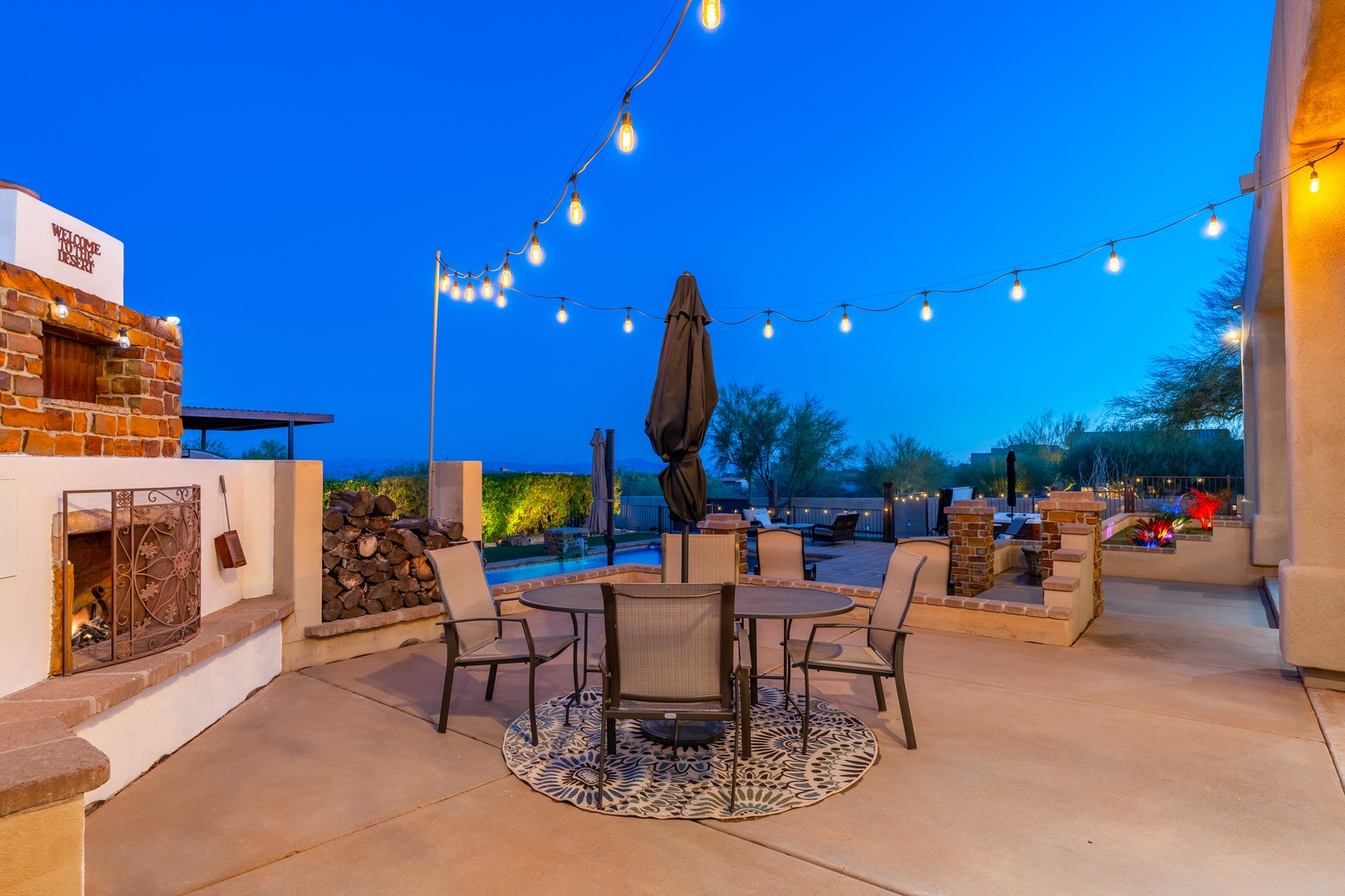 Evening scene showcasing the property's outdoor patio area, illuminated by string lights, with a fireplace, dining space, pool, and mountain backdrop under a twilight sky.