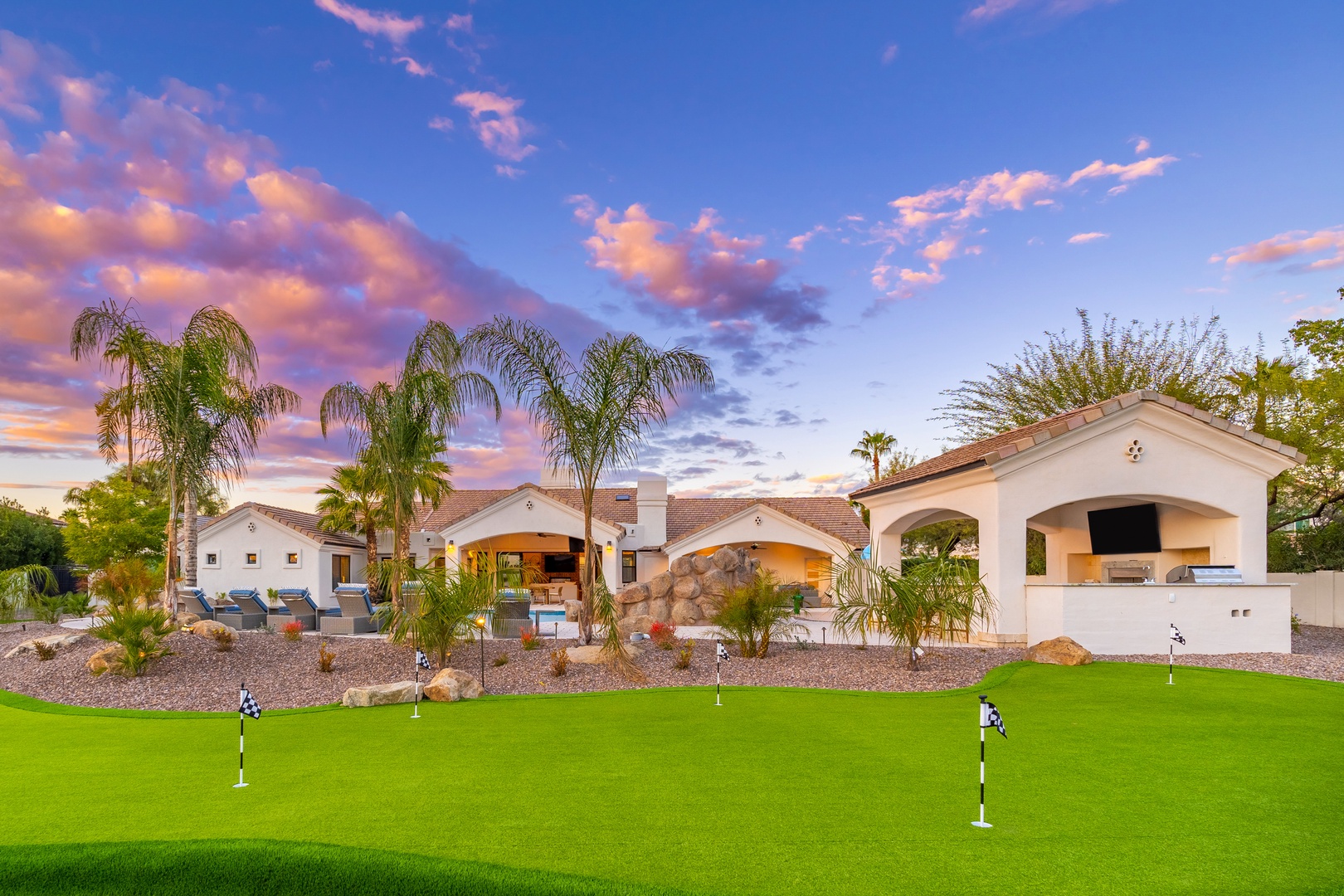 Stunning Spanish-style villa with outdoor entertainment pavilion, pristine putting green, and tropical palm landscaping captured at golden hour beneath vibrant desert skies.
