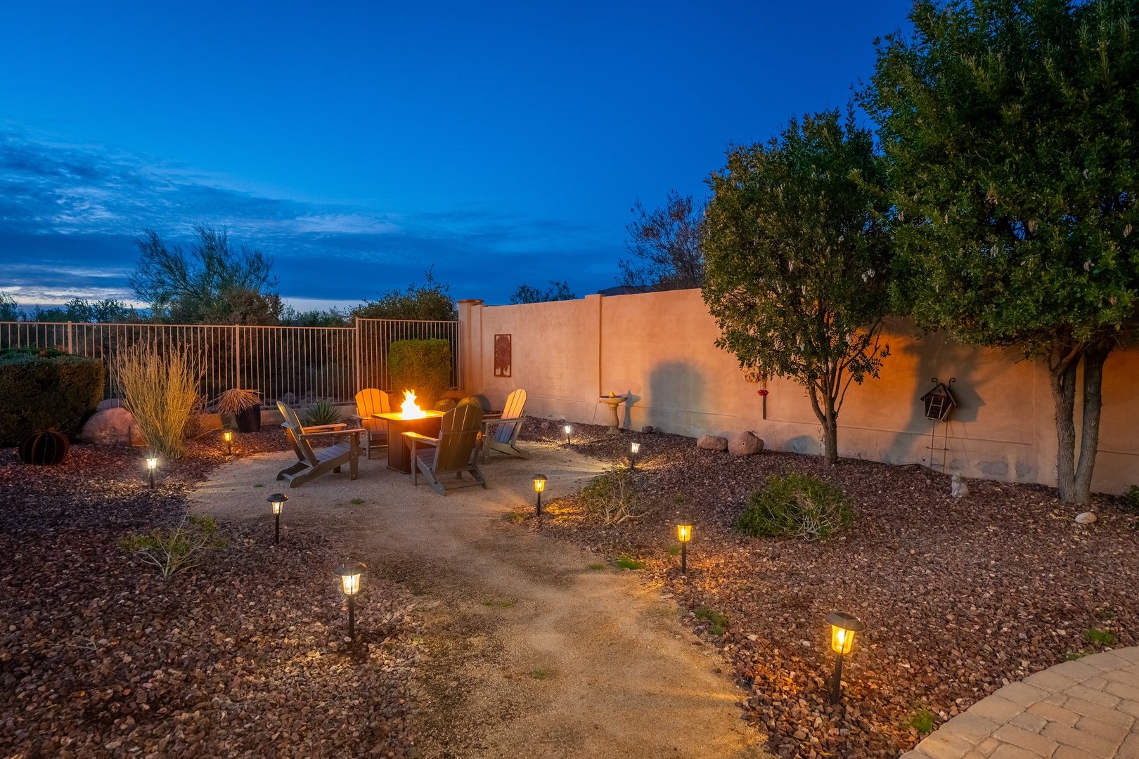 Twilight transforms the backyard into your private desert retreat, where a crackling fire pit, Adirondack chairs, and soft pathway lighting invite you to unwind under the stars.