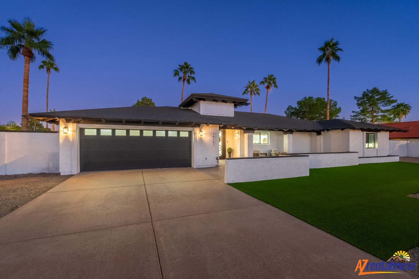 Modern single-story home with illuminated entrance and palm trees creating a warm desert oasis atmosphere at twilight.
