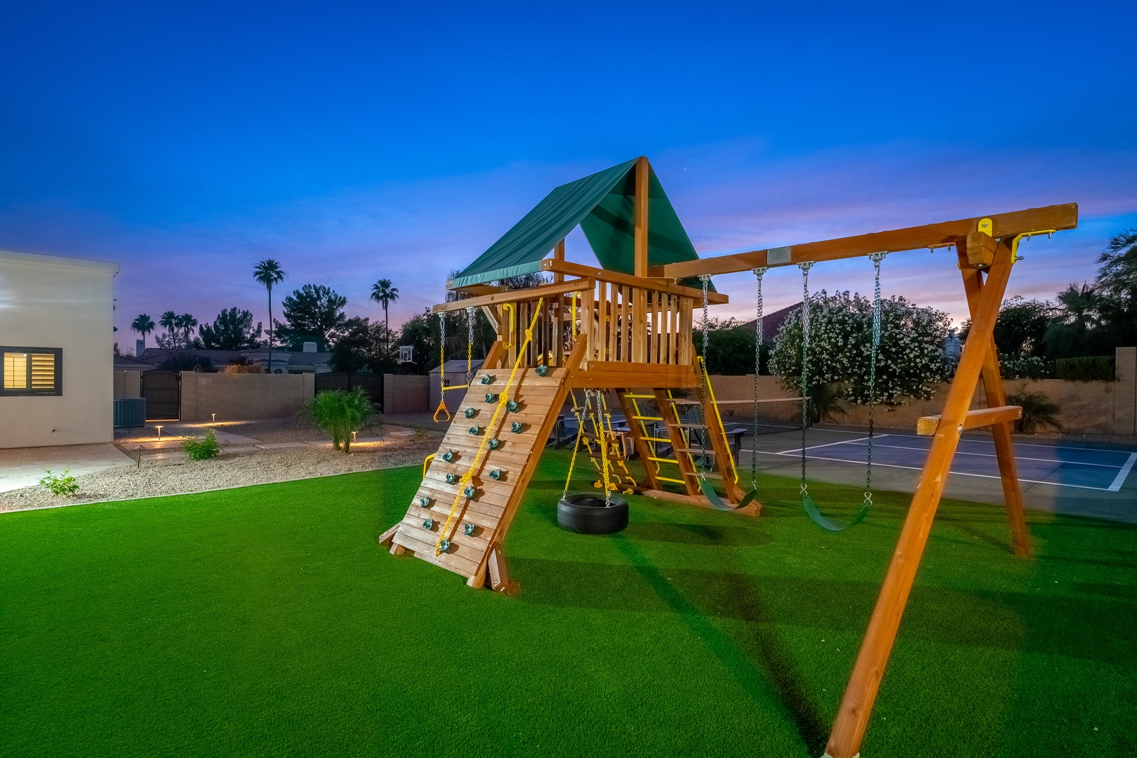A playground with swings, climbing wall, and covered platform stands ready on lush green lawn, with sports court and palm trees framing the backyard at dusk.