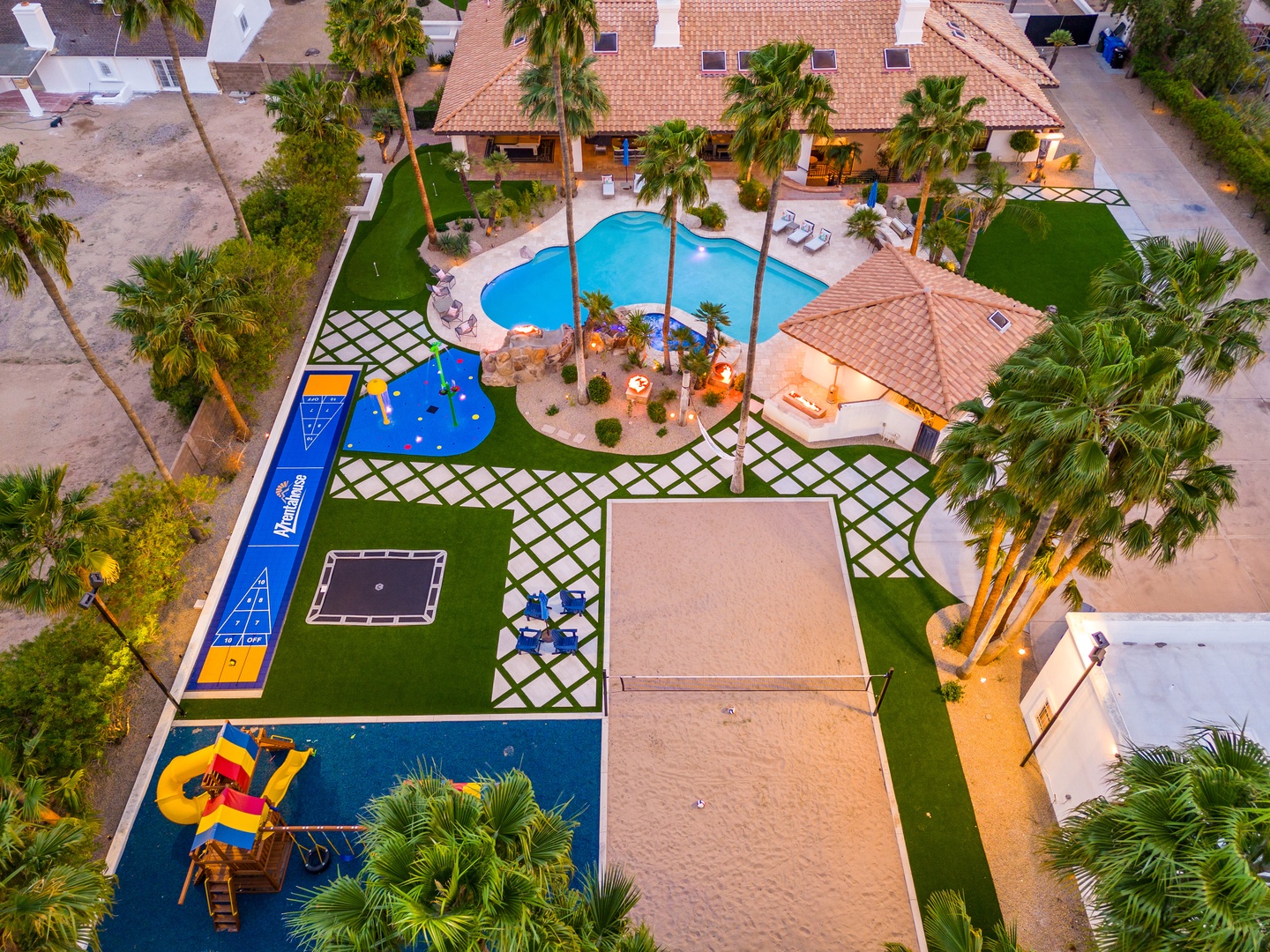 Aerial view of a resort-style vacation property featuring a sparkling swimming pool, children's play structures with water slides, spacious landscaped grounds with palm trees, outdoor recreation areas including shuffleboard, and multiple villa-style build