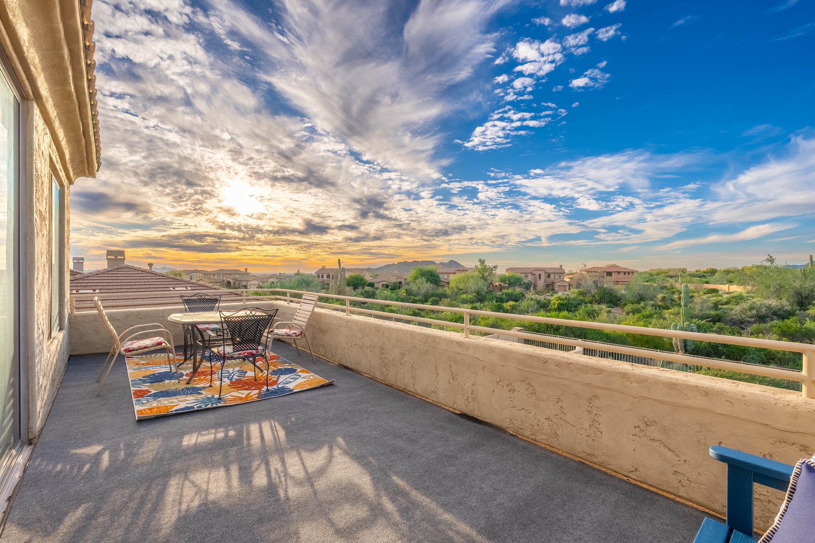 Expansive balcony overlooking a desert neighborhood with mountain views. Features outdoor seating beneath a dramatic sky, ideal for morning coffee or sunset evenings.