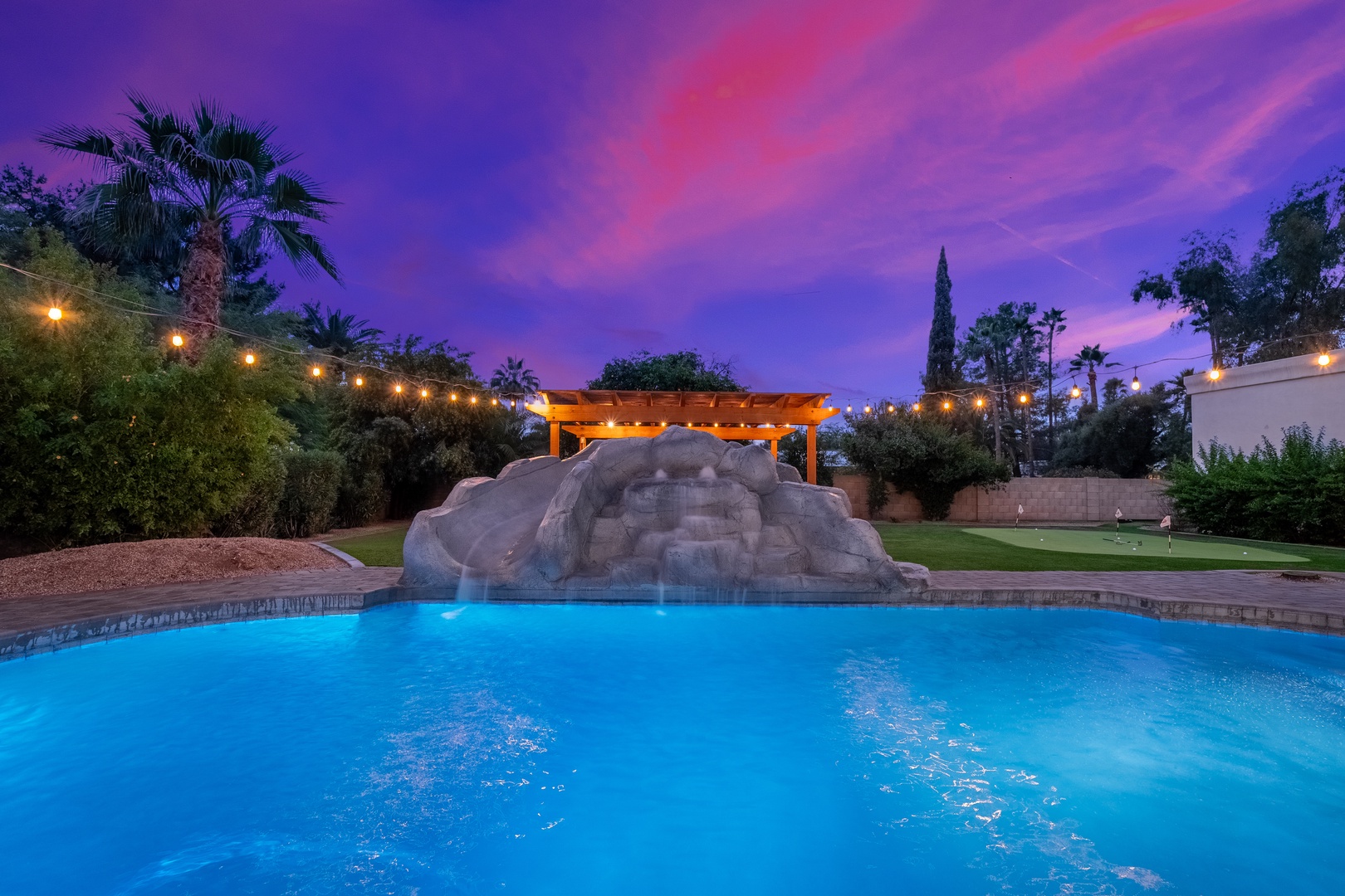 A stunning swimming pool with rock waterfall feature glows beneath a vibrant desert sunset, surrounded by lush palms and twinkling string lights—an enchanting evening escape.