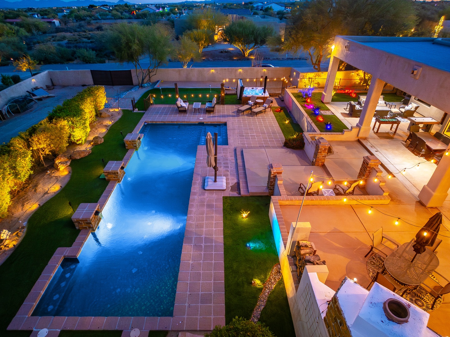 Aerial view of a luxury desert vacation property featuring a pristine swimming pool, multiple covered patios, lush landscaping, and ambient evening lighting throughout the expansive outdoor entertainment areas.
