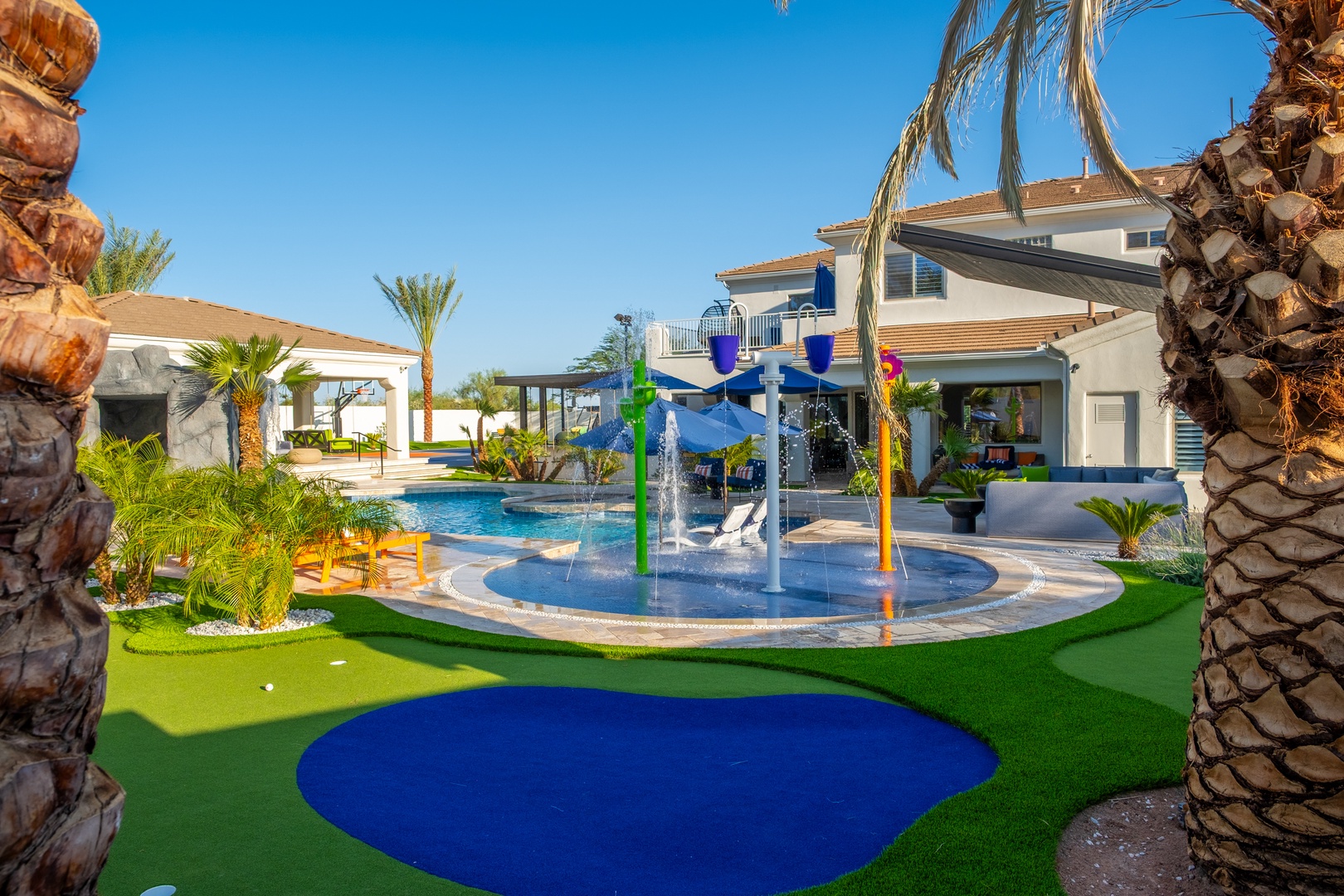 This stunning vacation villa features a vibrant splash pad, sparkling pool, and lush tropical landscaping under sunny blue skies—a perfect outdoor playground for your family's unforgettable getaway.