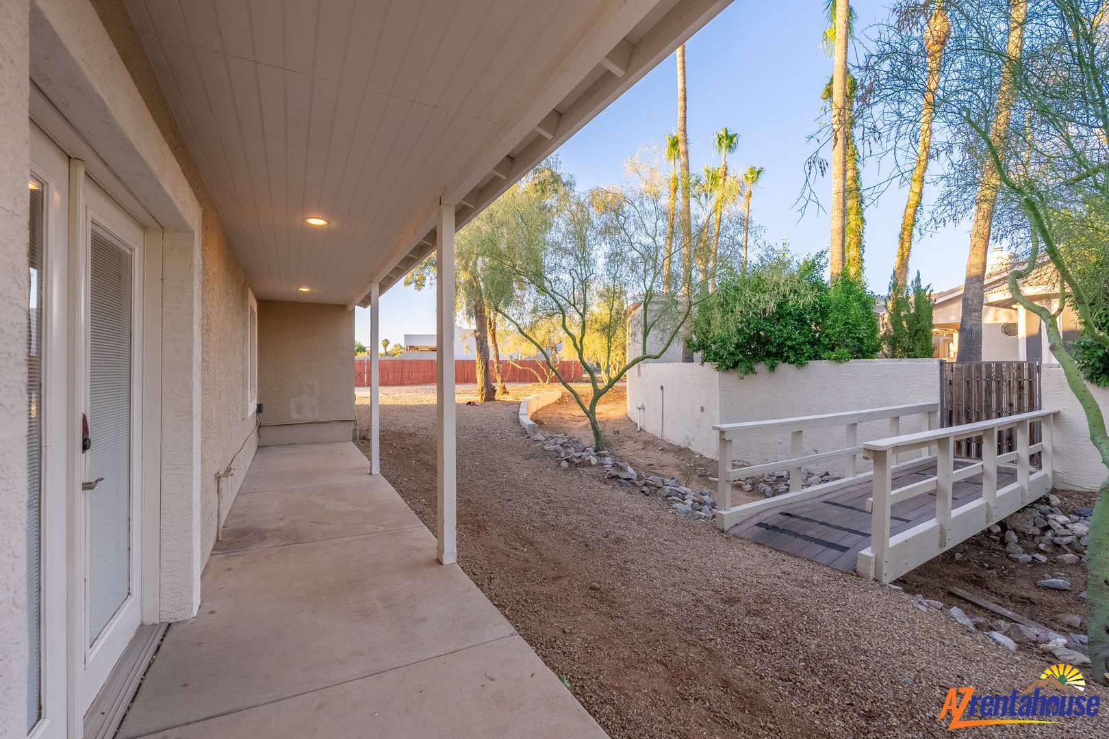 A peaceful side yard with a charming wooden footbridge over a decorative rock bed, framed by mature trees and tall palms that define the property's desert setting.