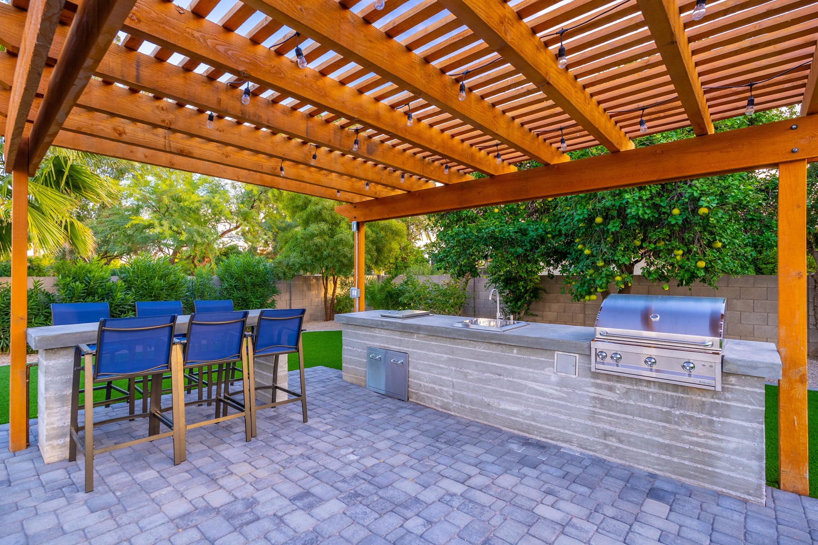 Gather under the pergola at your outdoor kitchen, featuring a built-in grill, ample counter space, and bar seating overlooking lush landscaping—perfect for entertaining in style.