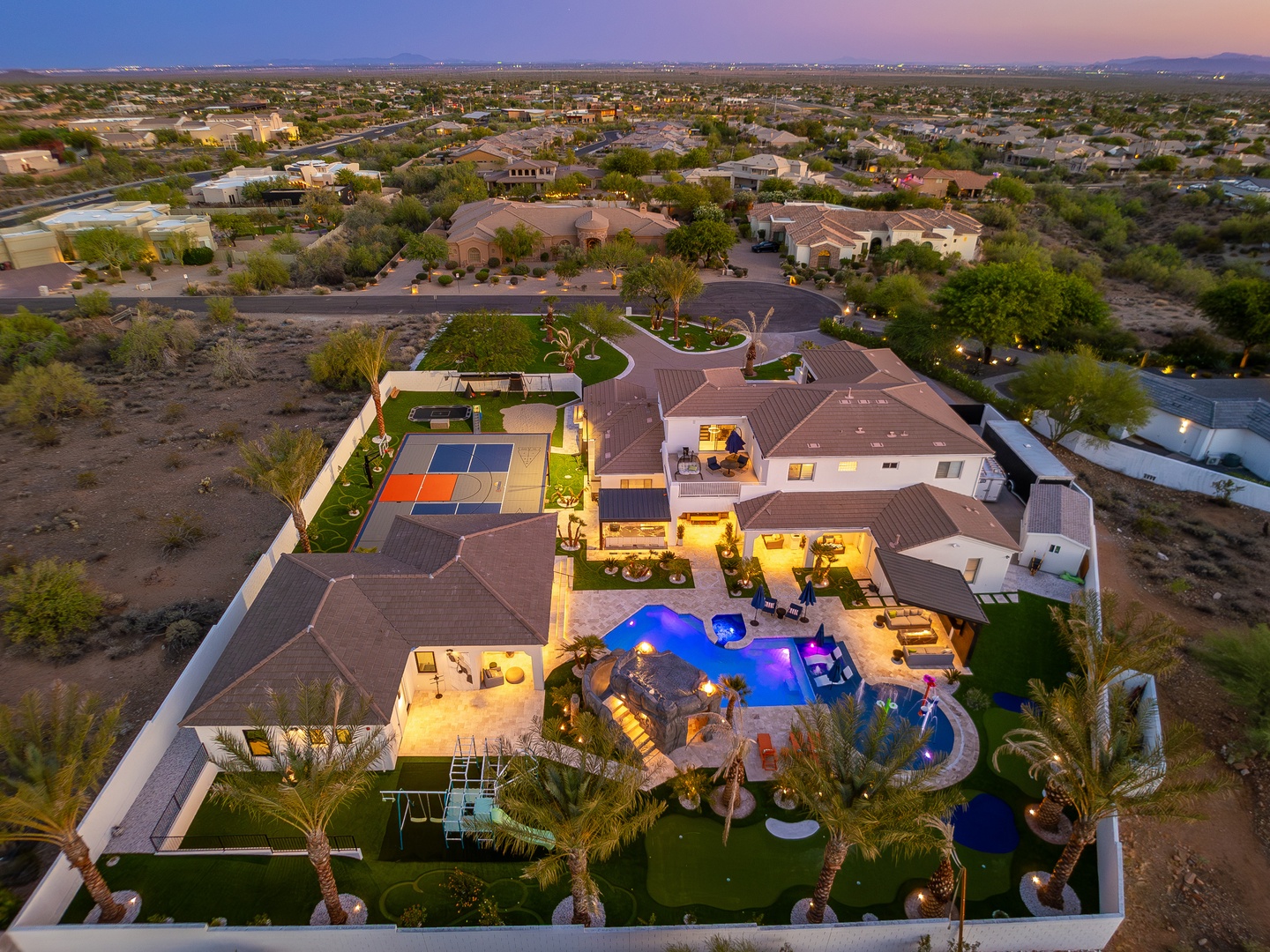 Aerial view of the property showcasing expansive resort-style grounds with pool, sports courts, and multiple outdoor entertainment areas set in a desert landscape at twilight.