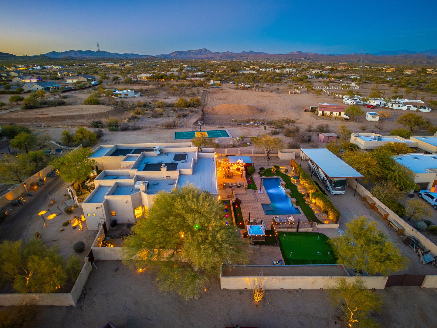 Aerial view of a desert vacation property featuring multiple pools, putting green, and outdoor entertainment areas, set against mountain vistas at dusk.