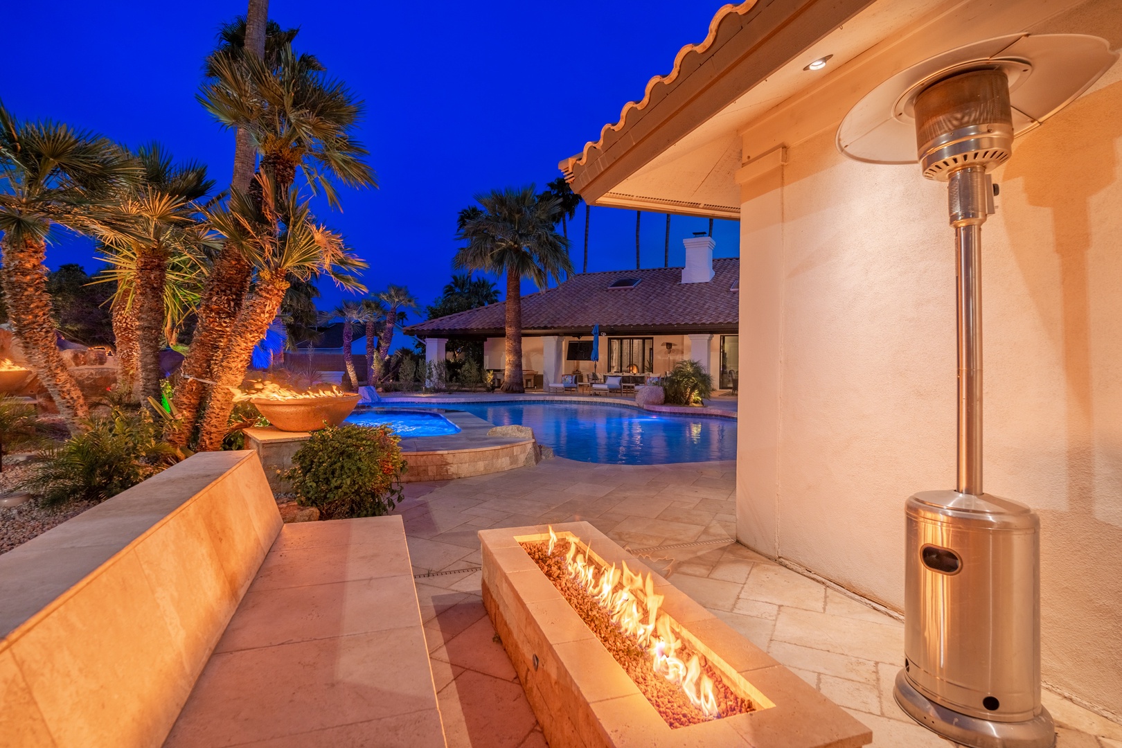 Stunning evening scene of the property featuring an illuminated pool, tropical palm trees, outdoor fire feature, and patio heater under a deep blue twilight sky.