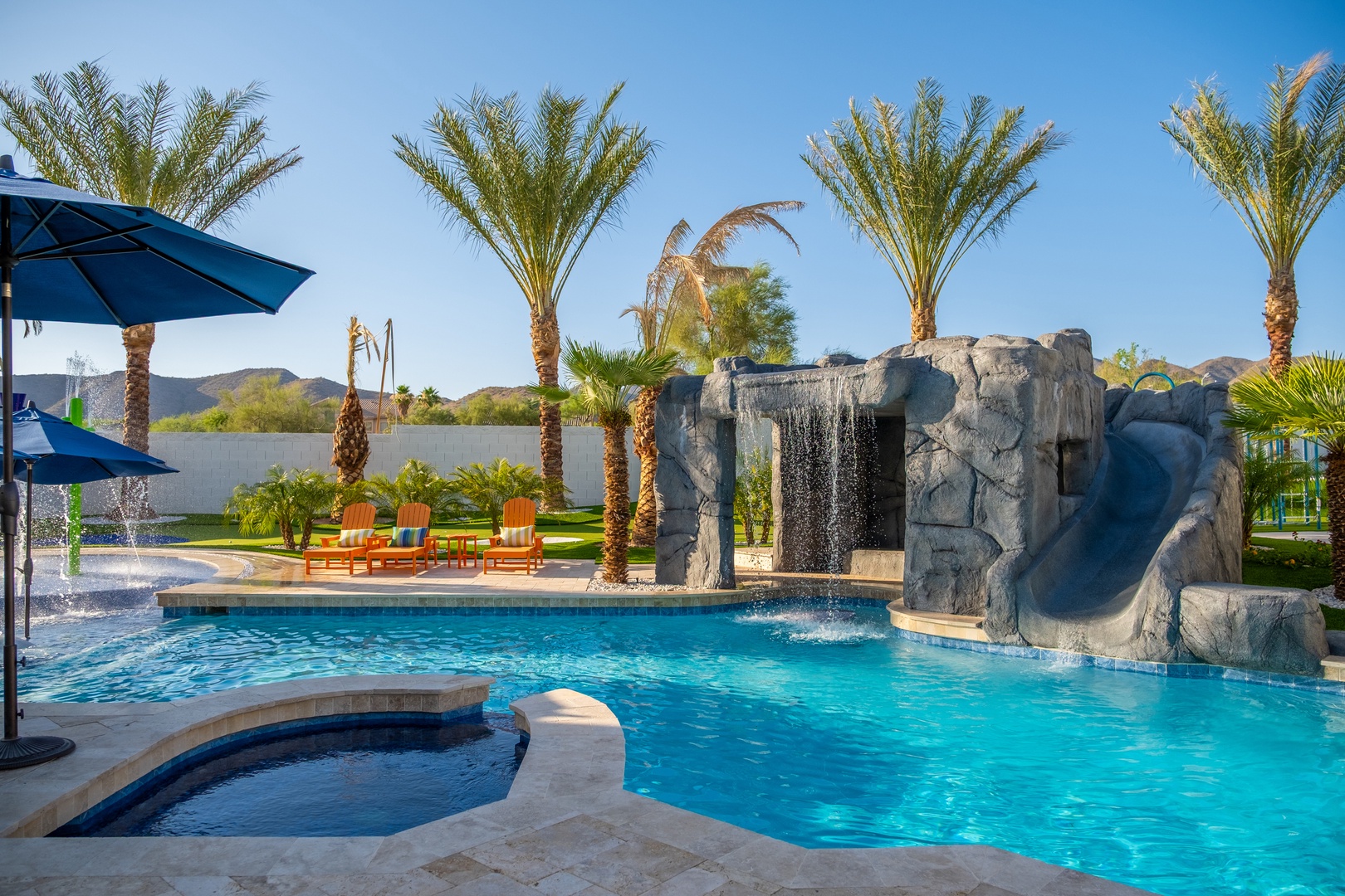 A resort-style pool features a dramatic rock waterfall and slide, surrounded by swaying palms and colorful lounge chairs, with mountain views framing the desert oasis.