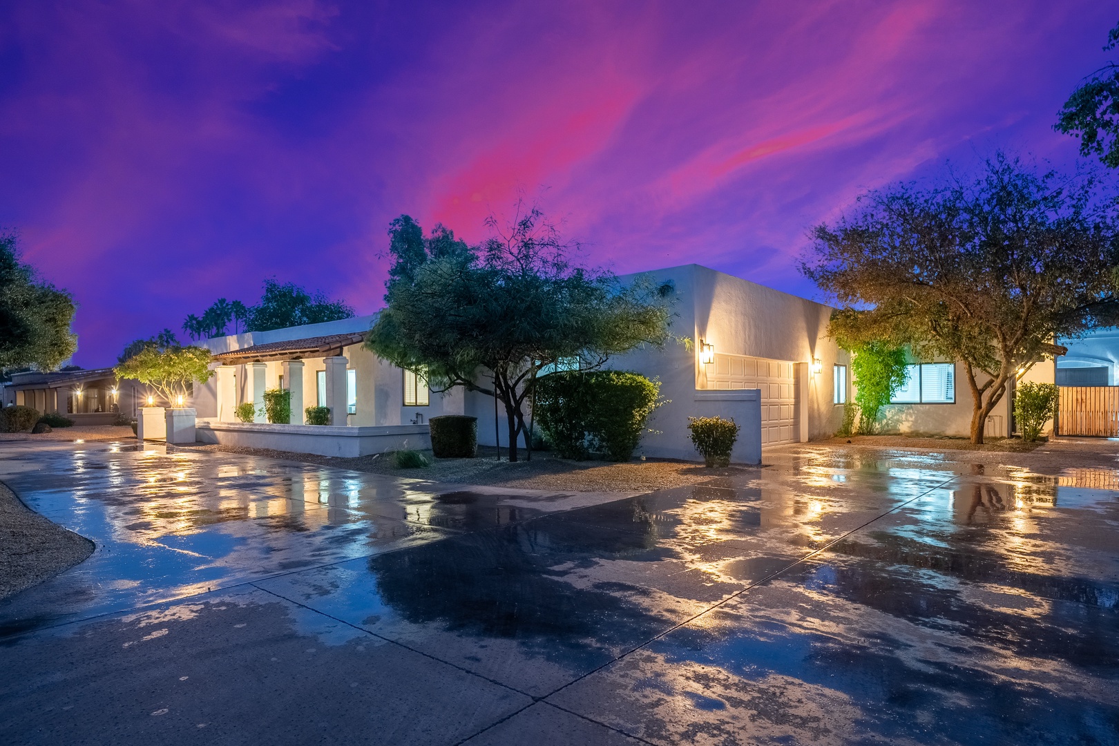 A stunning twilight arrival, as dramatic purple and pink skies frame the white stucco facade, landscaped trees, and exterior lighting that illuminate the property entrance and parking area.