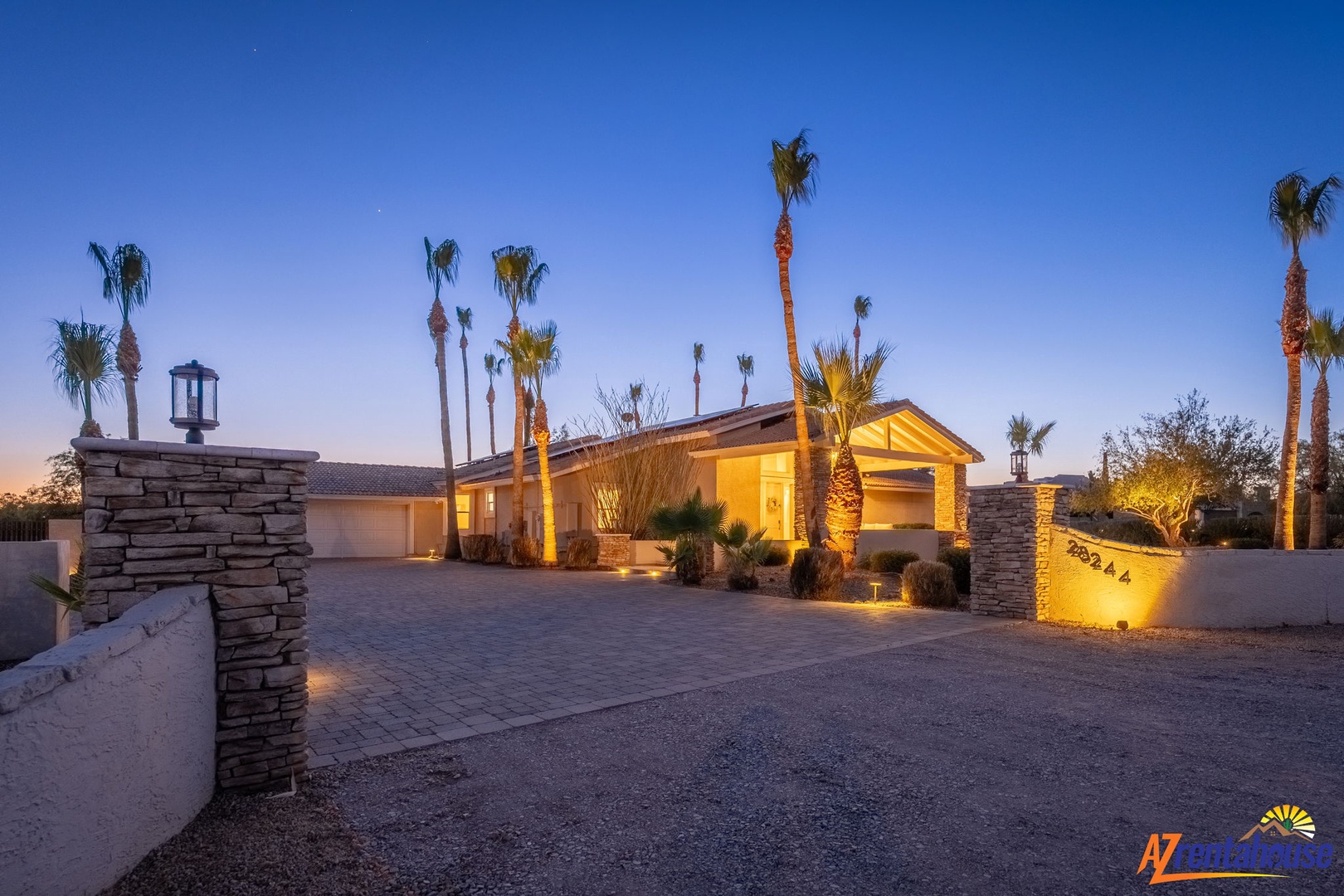 Desert property with stone-pillared entrance, illuminated ranch-style architecture, palm trees, and visible address marker at dusk.