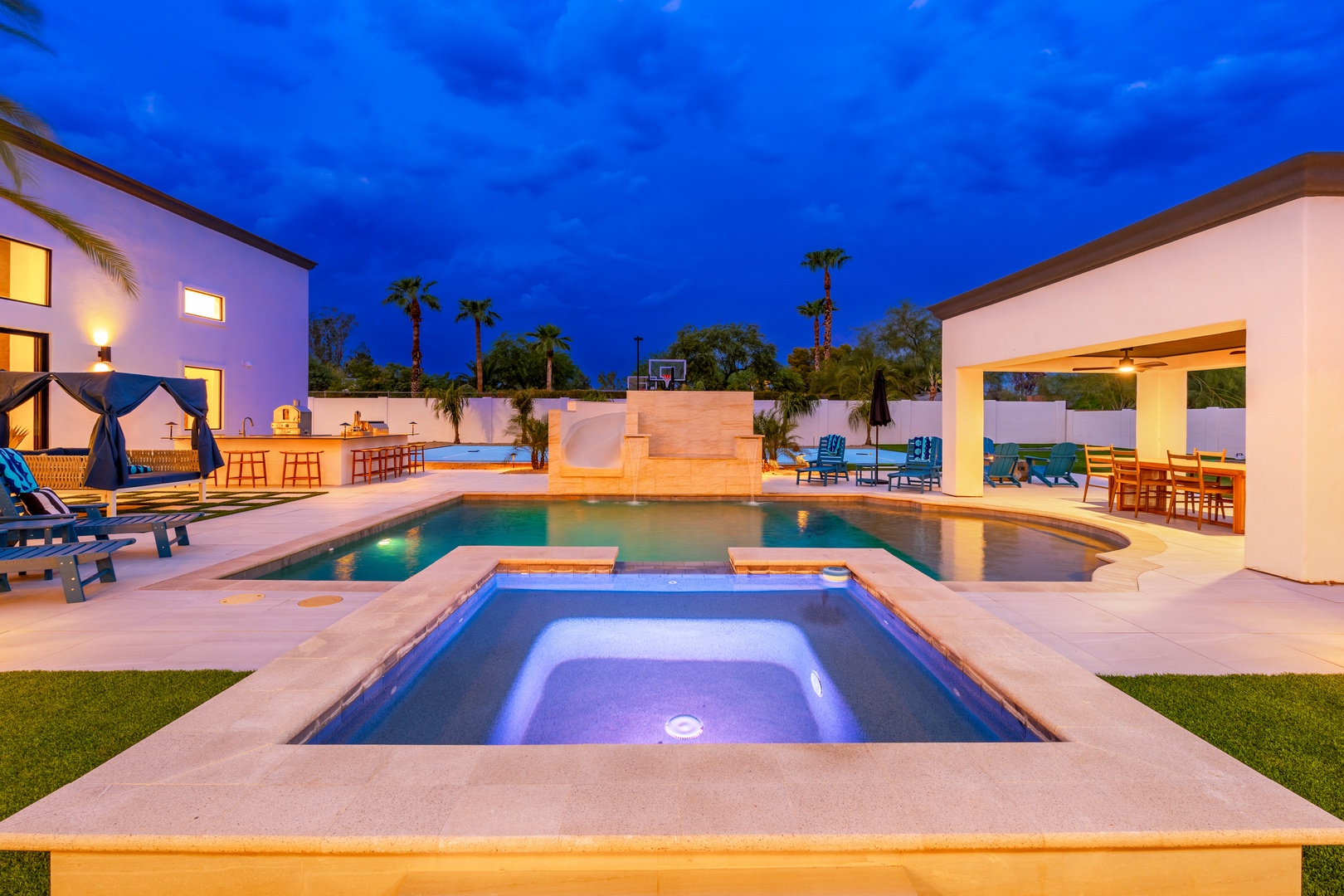 Modern desert oasis featuring a sparkling pool, illuminated spa, and stylish outdoor entertainment areas set against a stunning twilight sky with swaying palms.