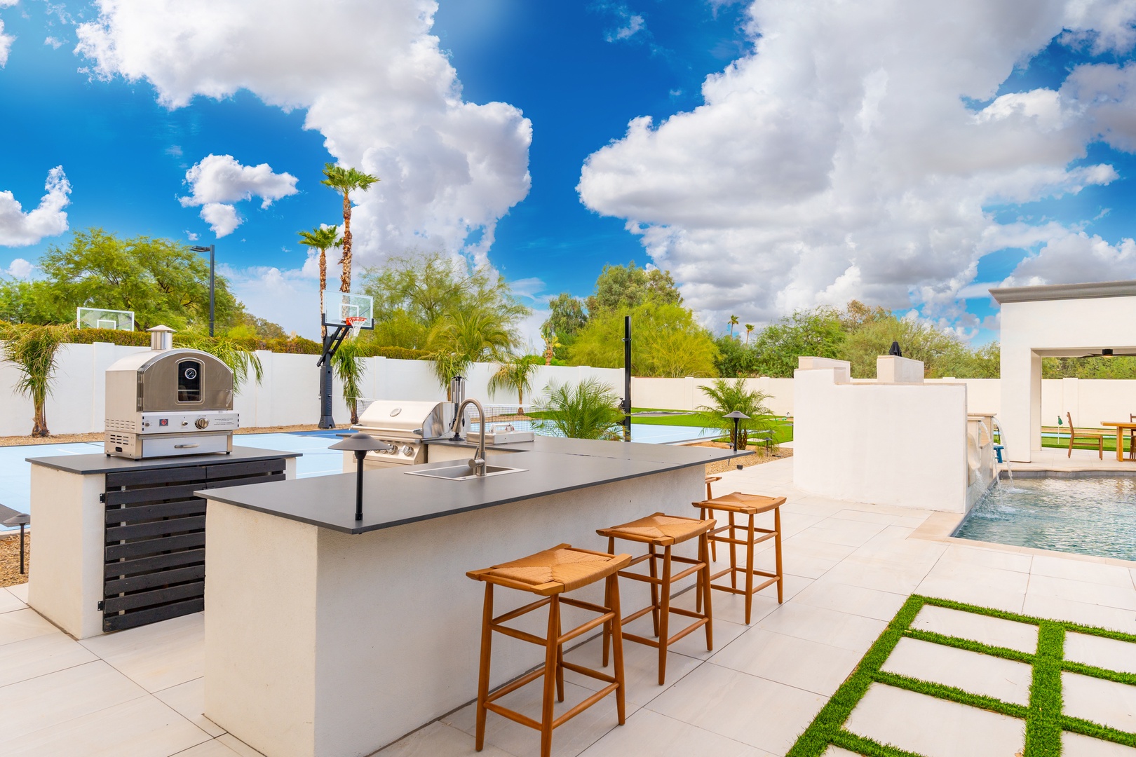 An outdoor oasis featuring a modern kitchen island with seating, built-in grills, lush tropical landscaping, a sparkling pool, and a basketball court under Arizona skies.