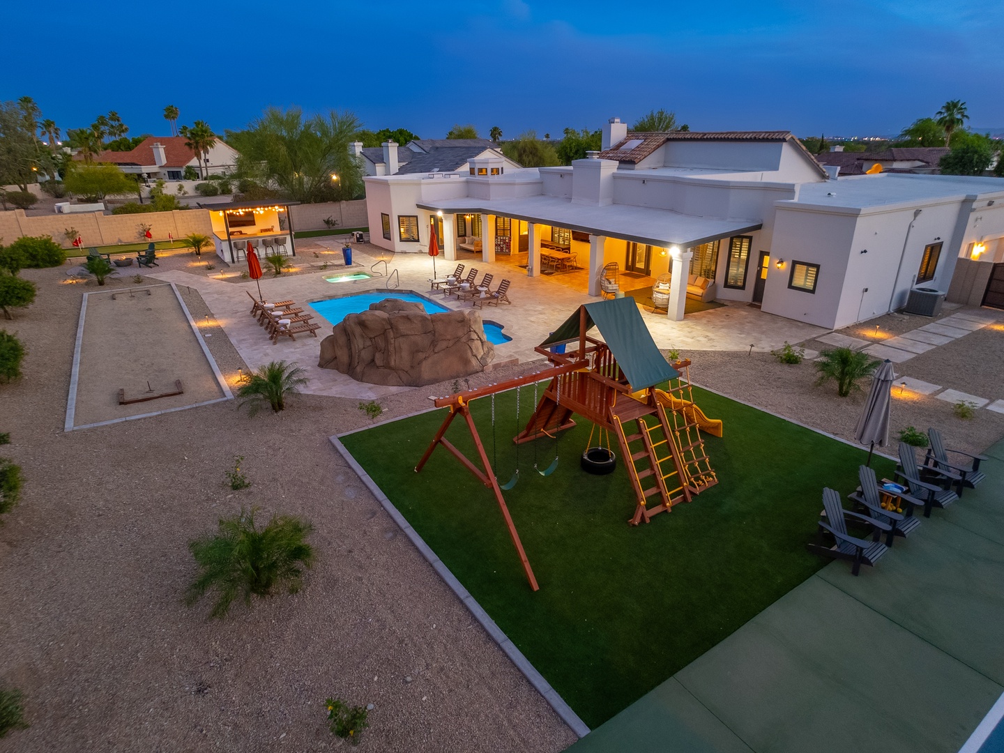 Aerial view of the property reveals a spacious backyard oasis with a sparkling pool, children's playground, covered patio, and luxurious outdoor living spaces beneath a desert twilight sky.