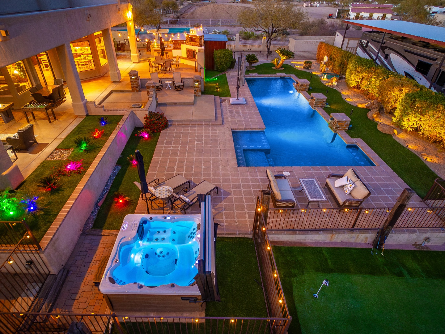 An aerial view of the beautifully lit backyard reveals a modern pool, hot tub, and spacious patio with colorful garden lighting perfect for evening entertaining and relaxation.