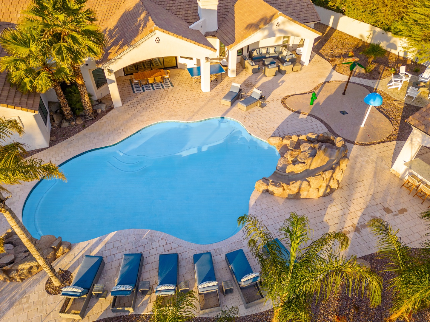 Aerial view of a luxury vacation property featuring a freeform pool, covered outdoor entertaining area, and lush tropical landscaping framed by palm trees.