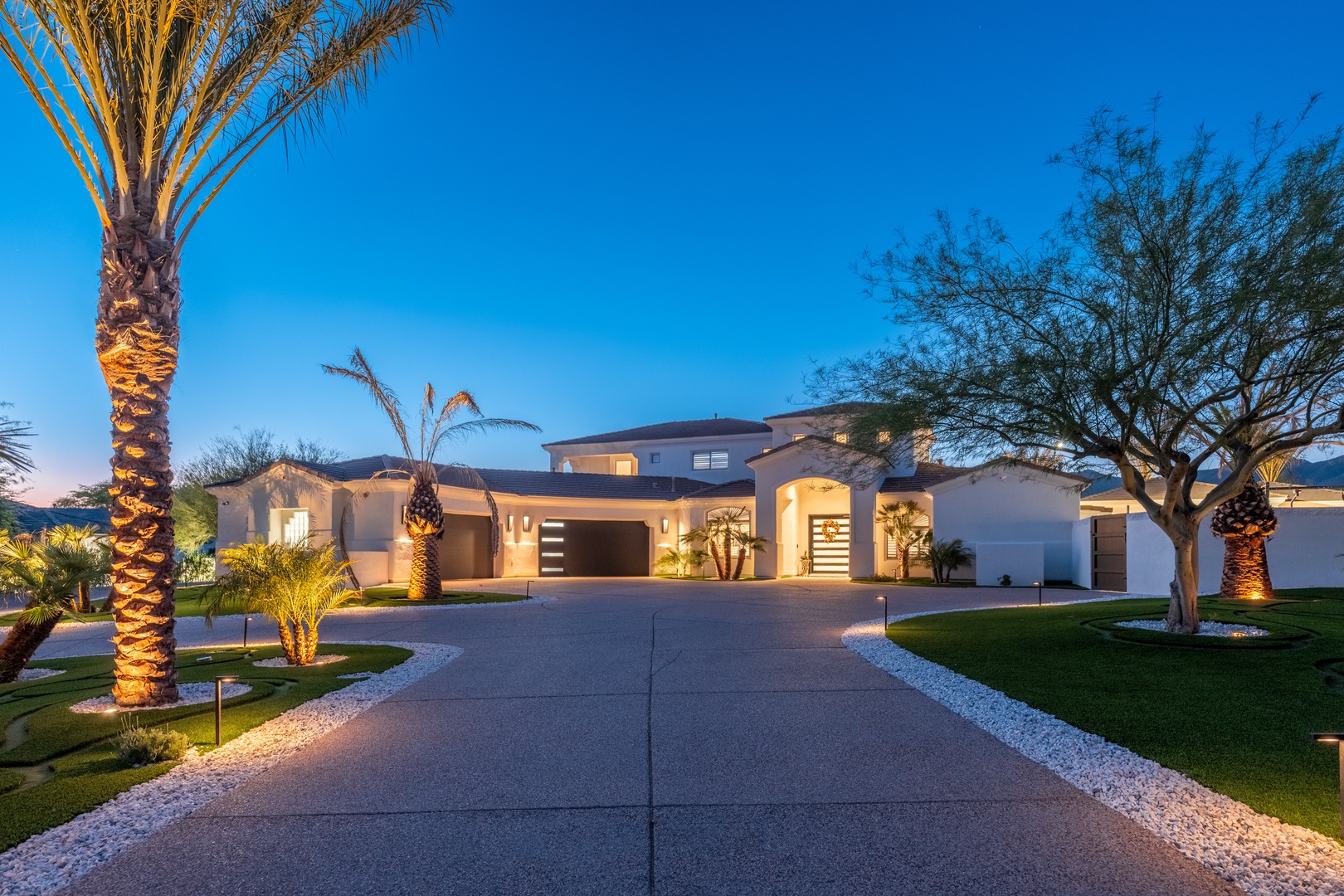 A grand modern property with illuminated palm trees and desert landscaping welcomes visitors along an expansive driveway as twilight settles over the surrounding area.
