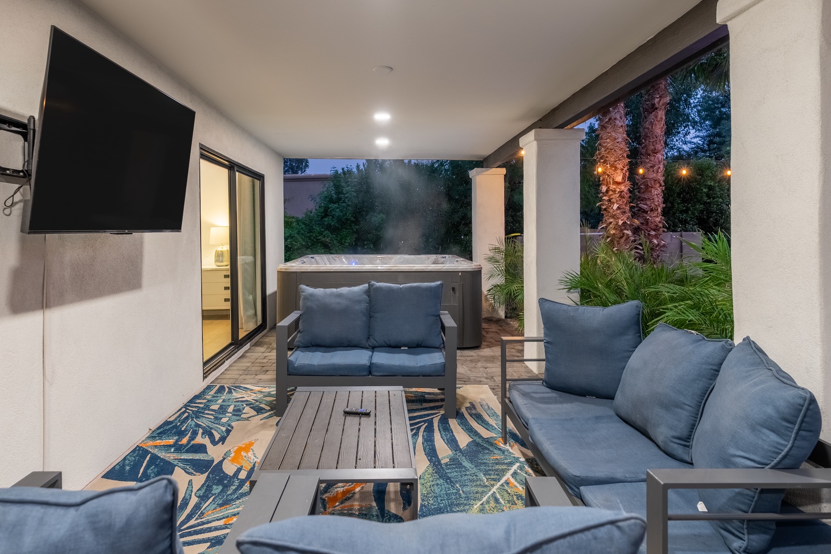 Picture yourself sinking into plush seating while your private hot tub bubbles nearby. This covered patio invites you to unwind under twinkling evening lights, with tropical palms swaying just beyond your retreat.