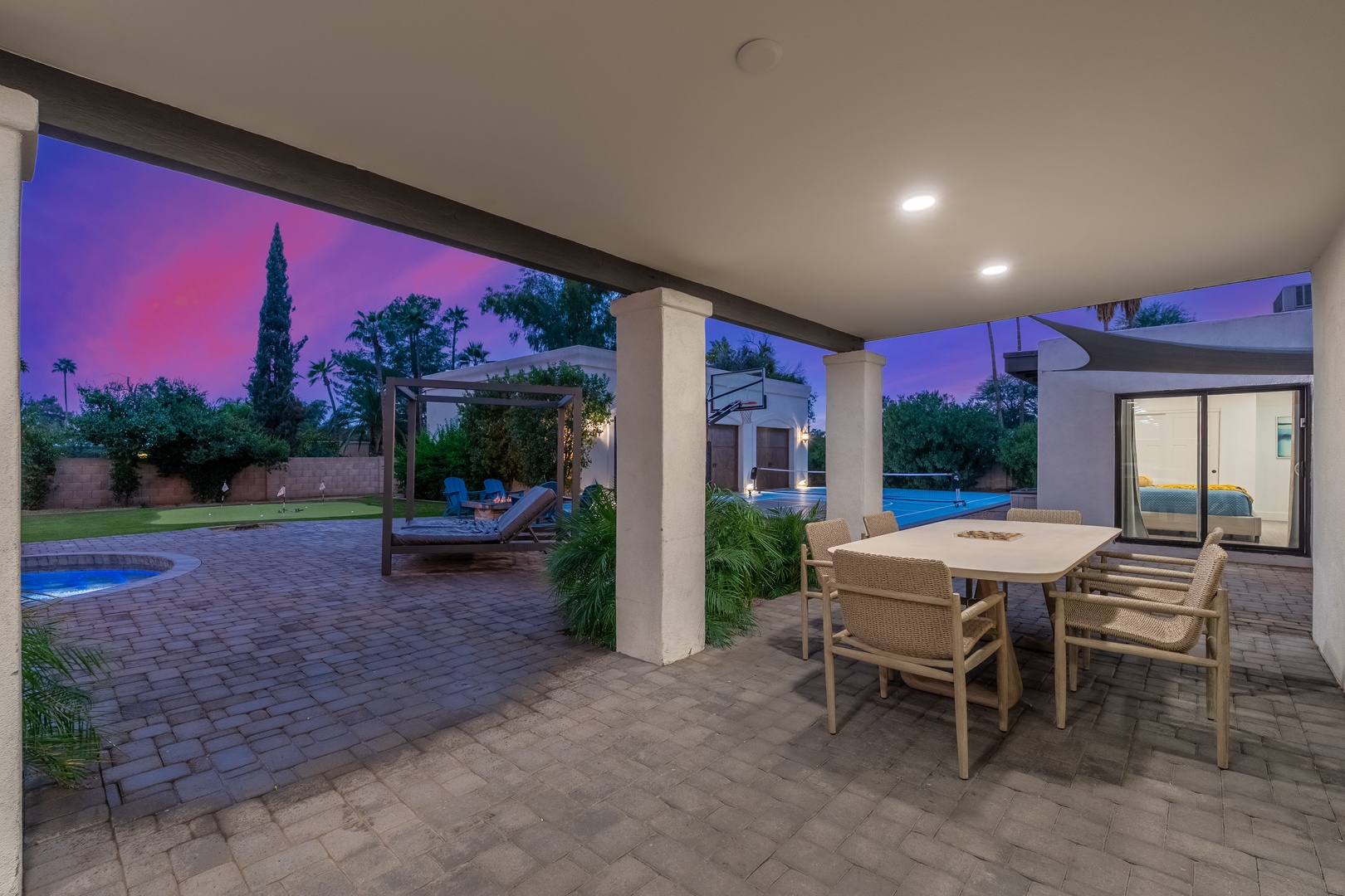 A stunning desert oasis awaits as twilight paints the sky in purple and pink hues. The covered patio offers prime seating for alfresco dining while the pool and loungers beckon just beyond.