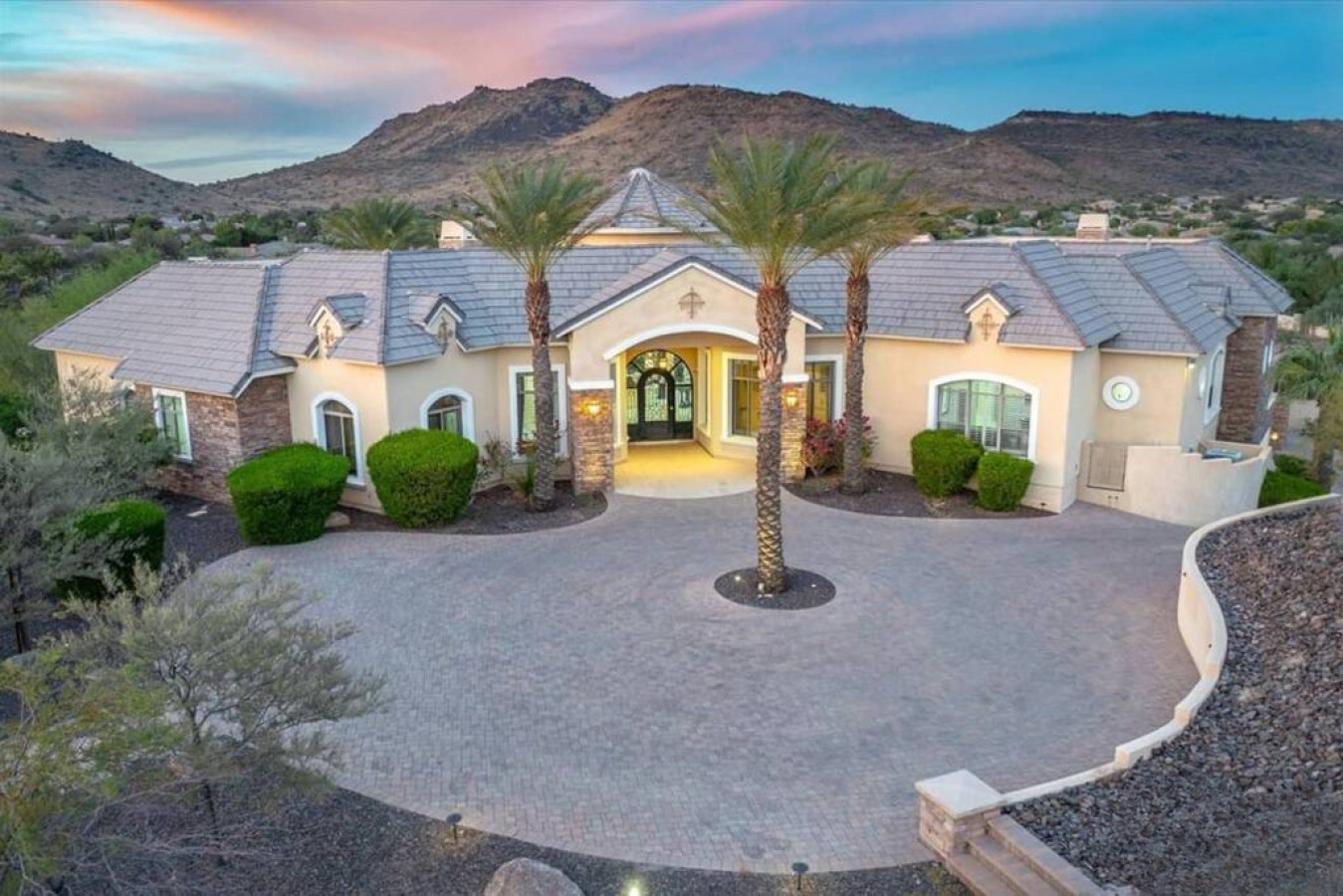 Elegant desert estate nestled against dramatic mountain vistas, featuring Mediterranean architecture, manicured landscaping, and private entry courtyard accessed via spacious paver driveway.