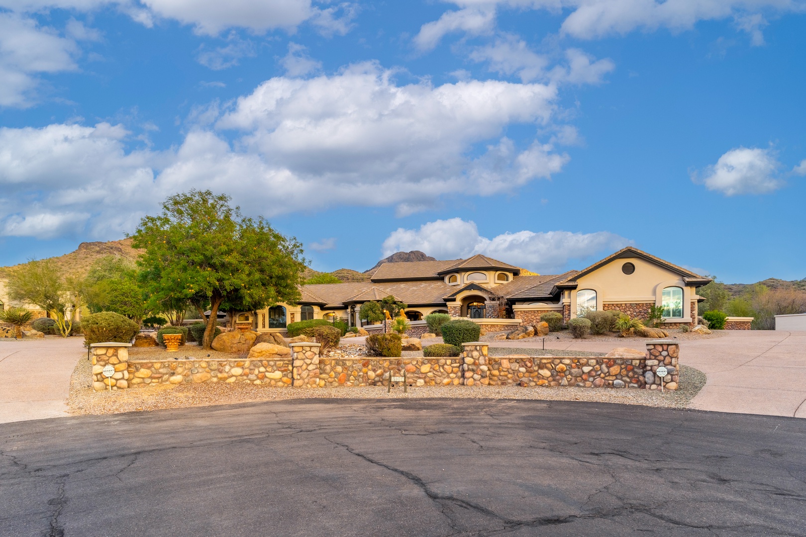 Stunning desert estate featuring Mediterranean-inspired architecture nestled against dramatic mountain vistas, framed by mature landscaping and distinctive stone-wall frontage welcoming travelers to the area.