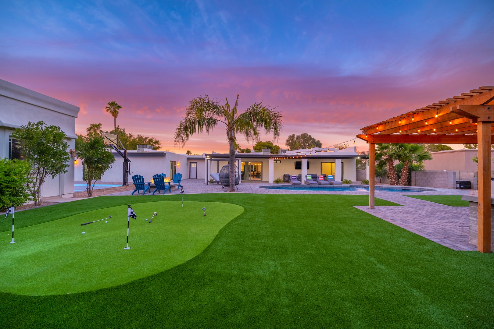 A stunning modern property featuring a private putting green, sparkling pool, and spacious outdoor entertaining areas beneath palm trees and a breathtaking painted sky.